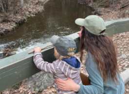A woman with long brown hair in a green ballcap kneels next to a little boy in a blue ballcap and striped hoodie next to a stream surrounded by brown fallen leaves