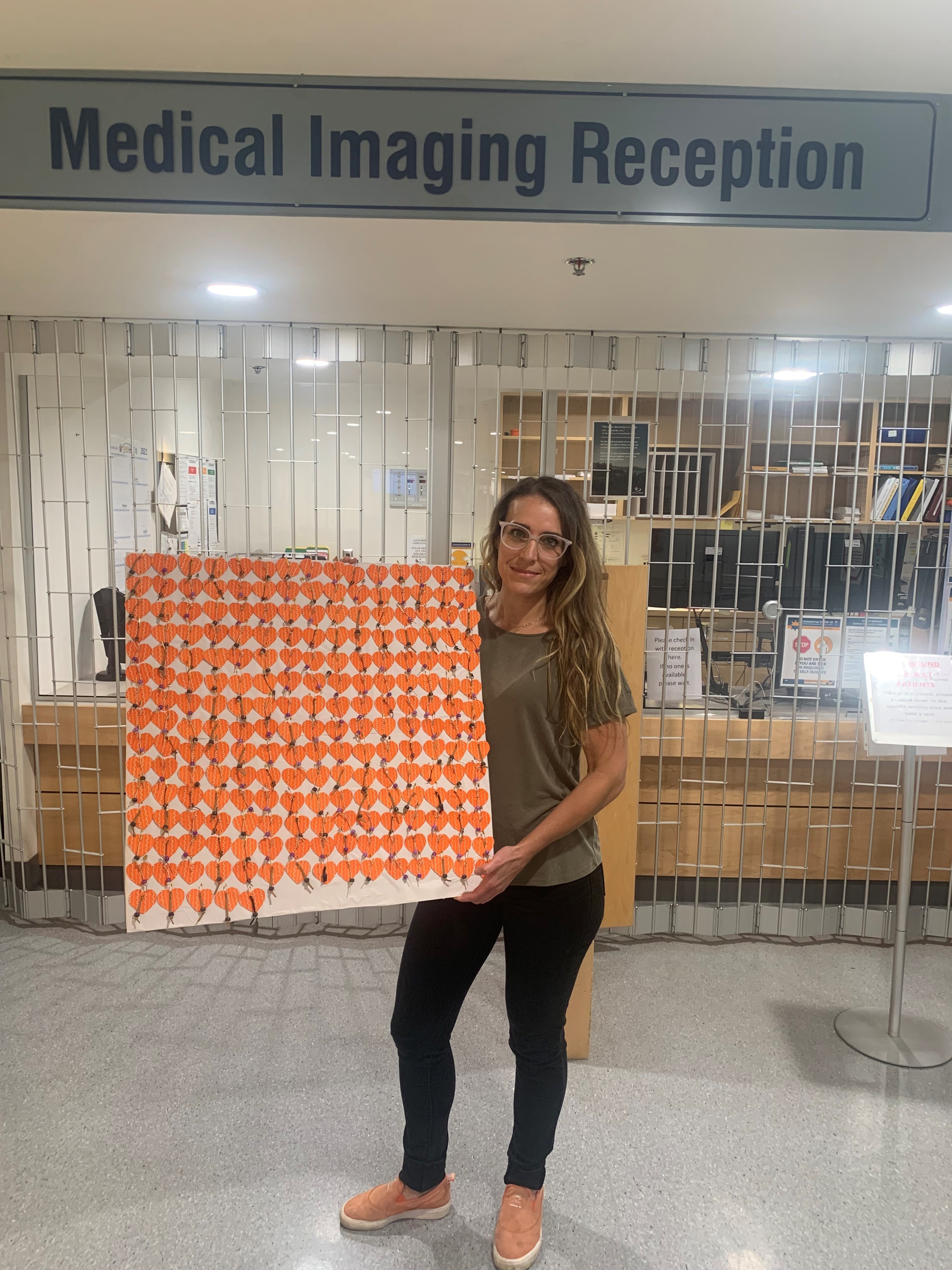 A long haired brunette wearing glasses holds up a poster with many small orange hearts on it in the reception area of the Medical Imaging department at the hospital 