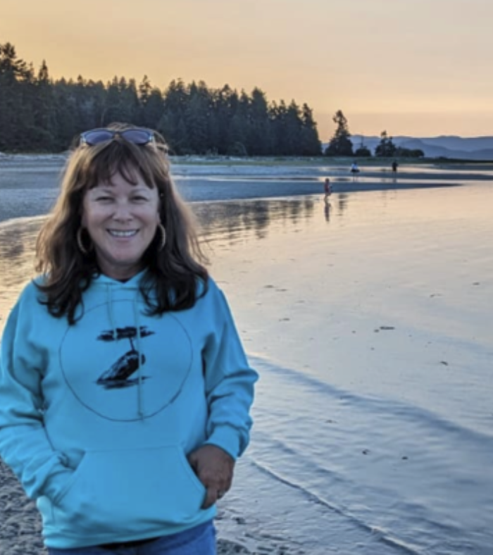 a smiling brunette woman stands with hands in her hoody pocket on the beach right next to the water