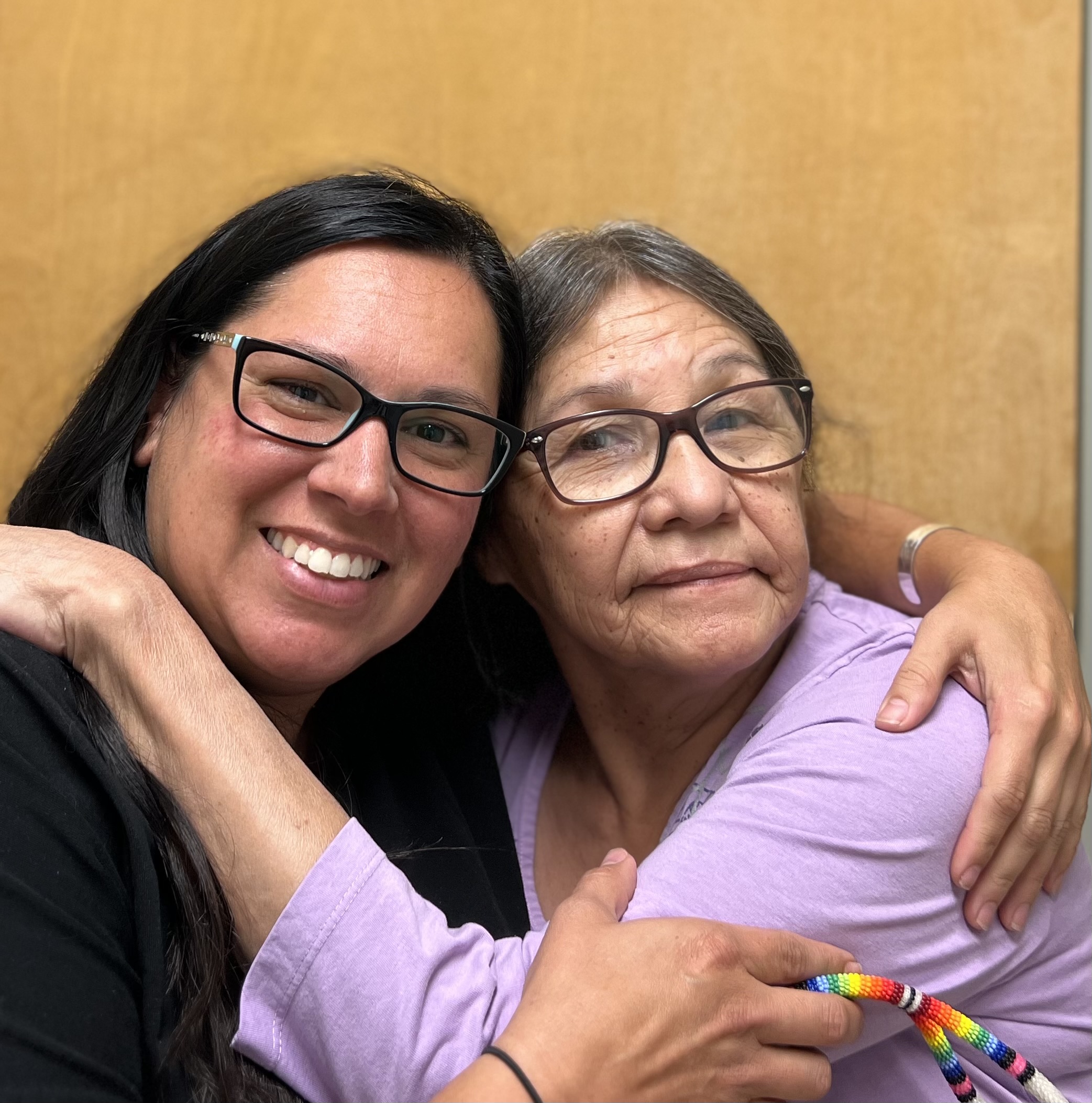 An Indigenous woman with hair in braid hugs an Indigenous woman with brown hair and glasses  