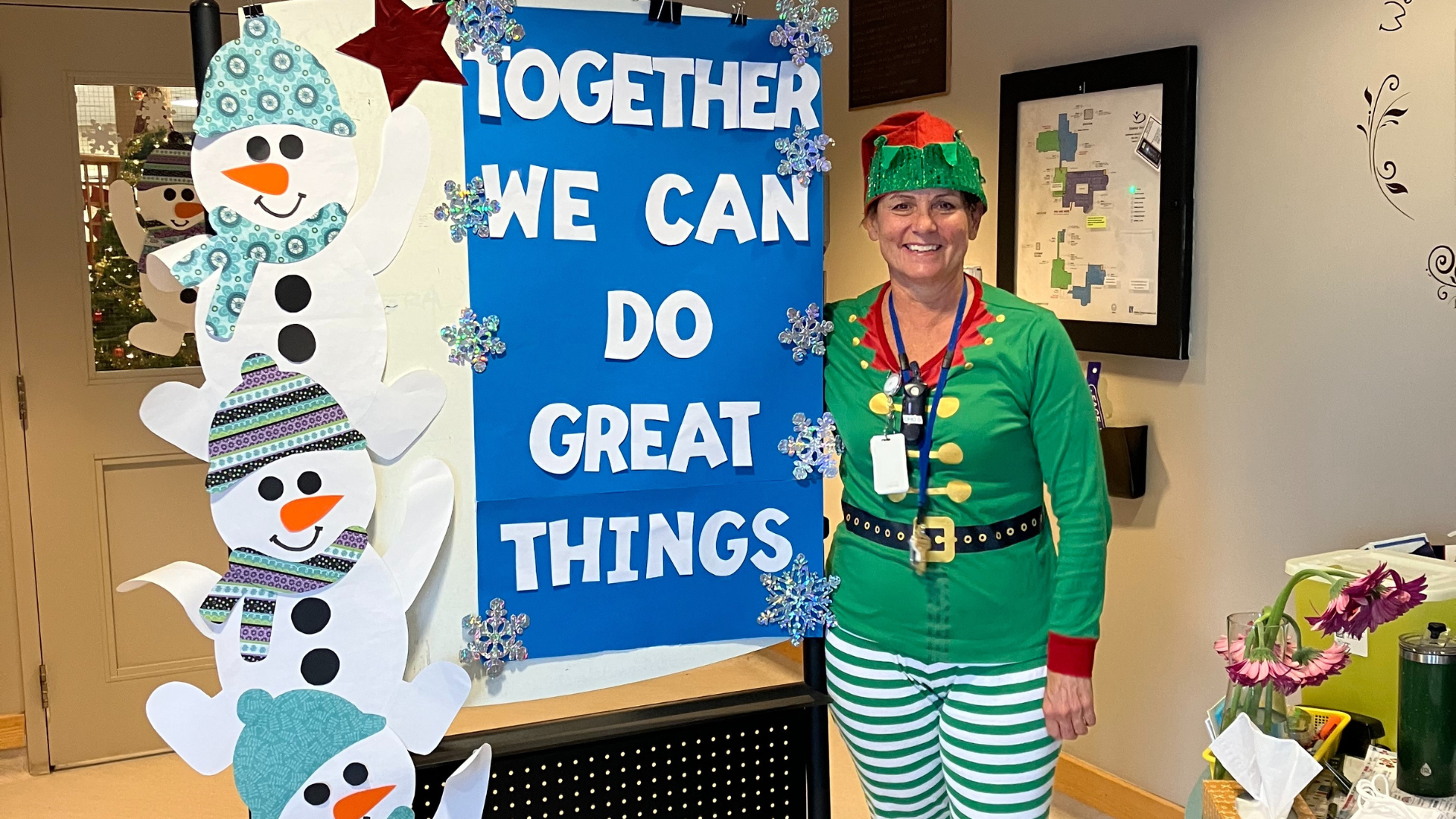 A woman dressed as an elf standing next to a blue Christmas sign that reads: “together we can do great things.”