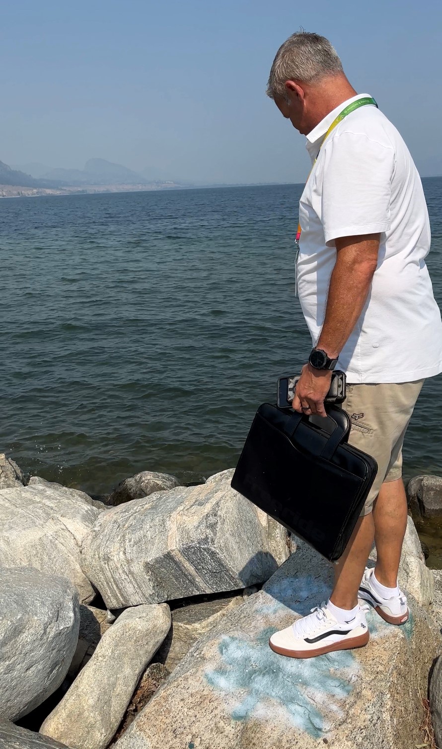 A man in a white tshirt and beige shorts carrying a black briefcase stands on a rock in front of a lake and points down