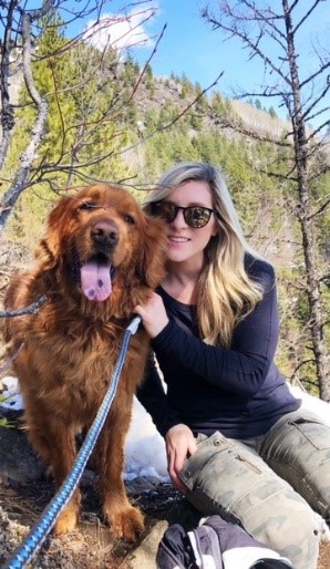 A woman with long blonde hair wearing sunglasses, a black long-sleeved shirt and camouflage pants sits with her red golden retriever on a hiking trail with a mountain in the background