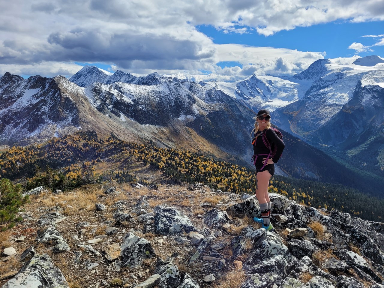 A woman with blonde pigtails wearing a black ball cap, pullover and shorts and purple backpack stands with her hands on her hips on a mountain peak set against a field of yellow flowers and snowcapped mountains