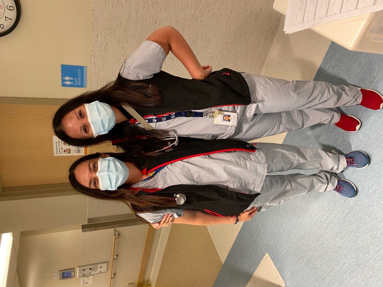 Two women stand side by side with their arms around each other's shoulders in a hospital hallway. Both are wearing grey uniforms, black vests, face masks and stethoscopes