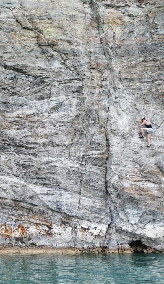 A zoomed out view of a woman climbing a huge rock face above water