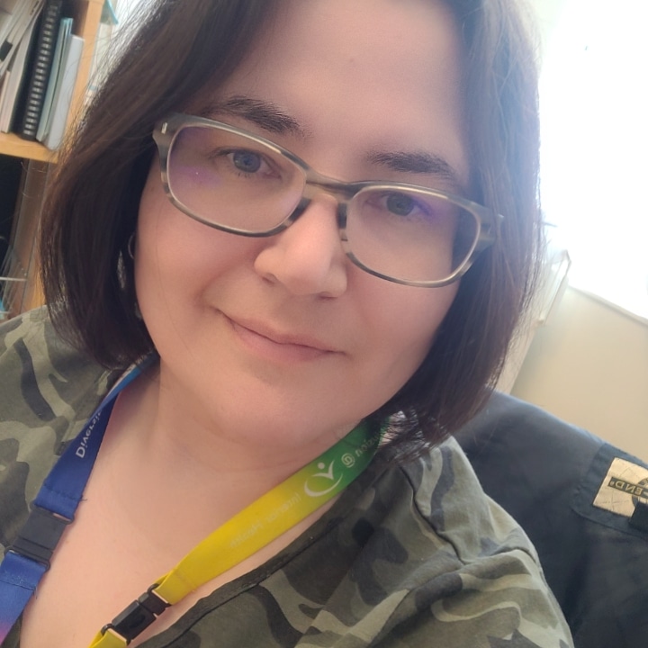 A woman with brown medium-length hair and glasses and wearing a rainbow lanyard and green camouflage shirt sits in a grey chair