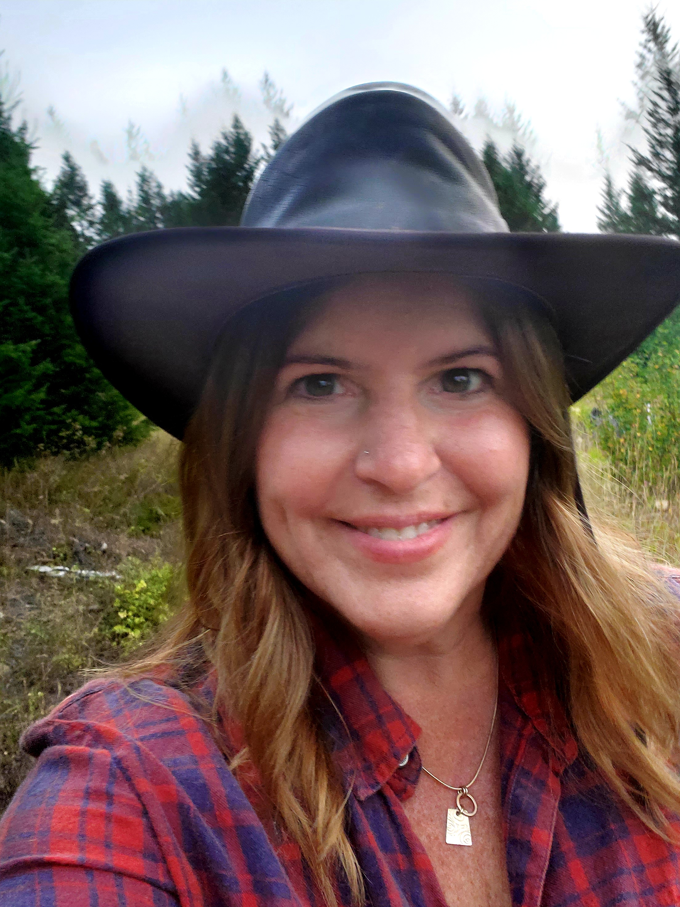 A woman with light brown hair wearing a black cowboy and a plaid shirt smiling at the camera