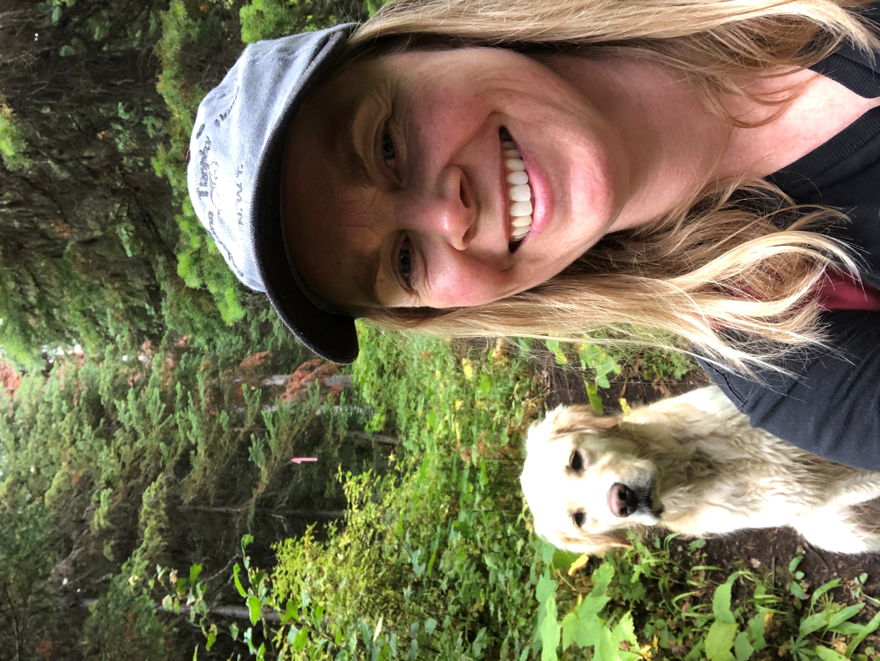 A smiling woman with long blond hair in a blue ball cap takes a selfie in a forest with her cream coloured dog behind her.