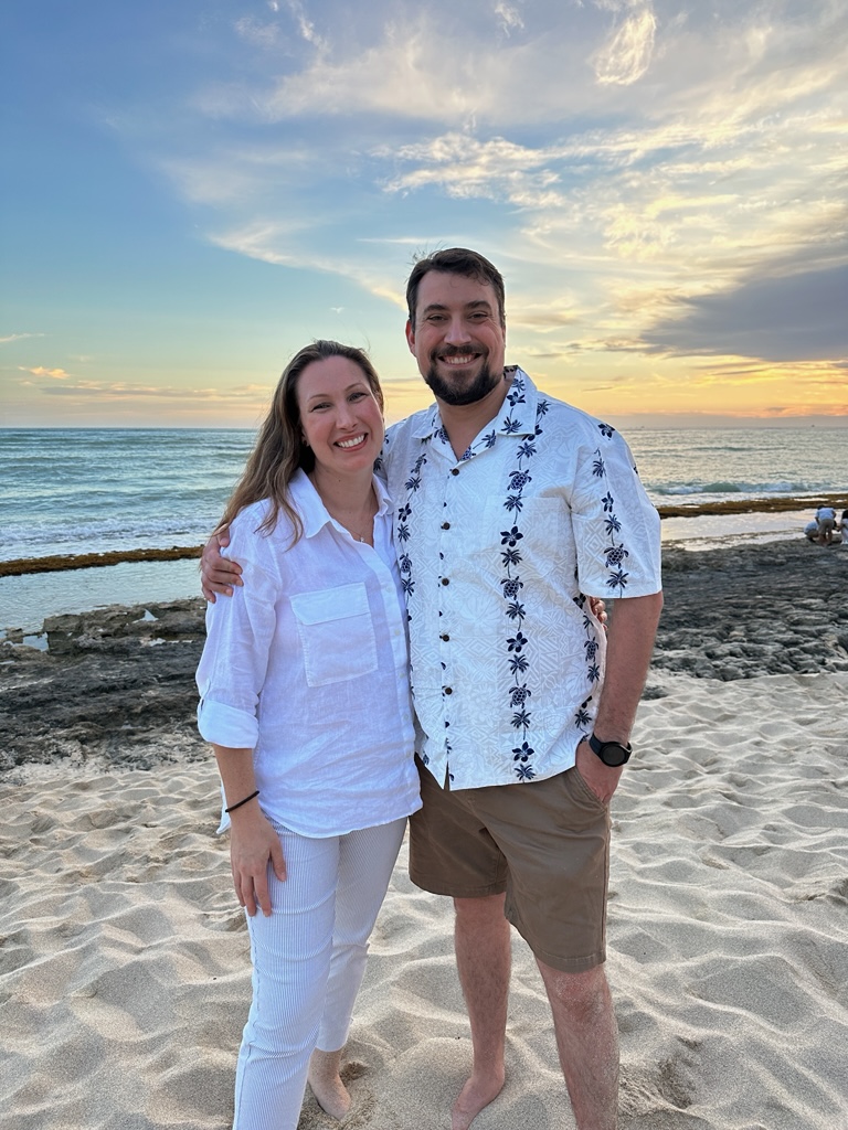 A man with dark hair and facial hair, has his arm around a woman with long brown hair. They stand on a beach in front of the ocean, with a sunset in the background. She wears a white button-up and white pants, he wears a white Hawaiian shirt with khaki shorts and a black wristwatch. 