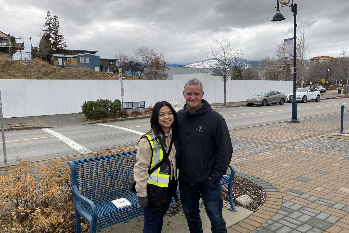 A woman with long dark hair wearing a safety vest and beige coat stands next to a man with a black jacket and blue jeans in front of a blue park bench on a cobbled street with snowy mountains behind them