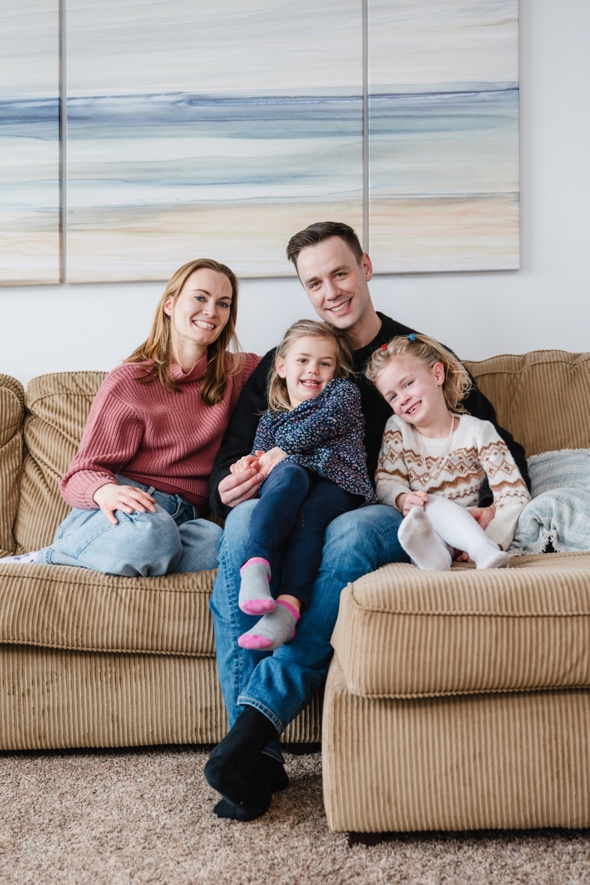 A mom in a pink sweatshirt, a dad in jeans and two toddlers sit on a beige couch in front of a landscape painting in a living room