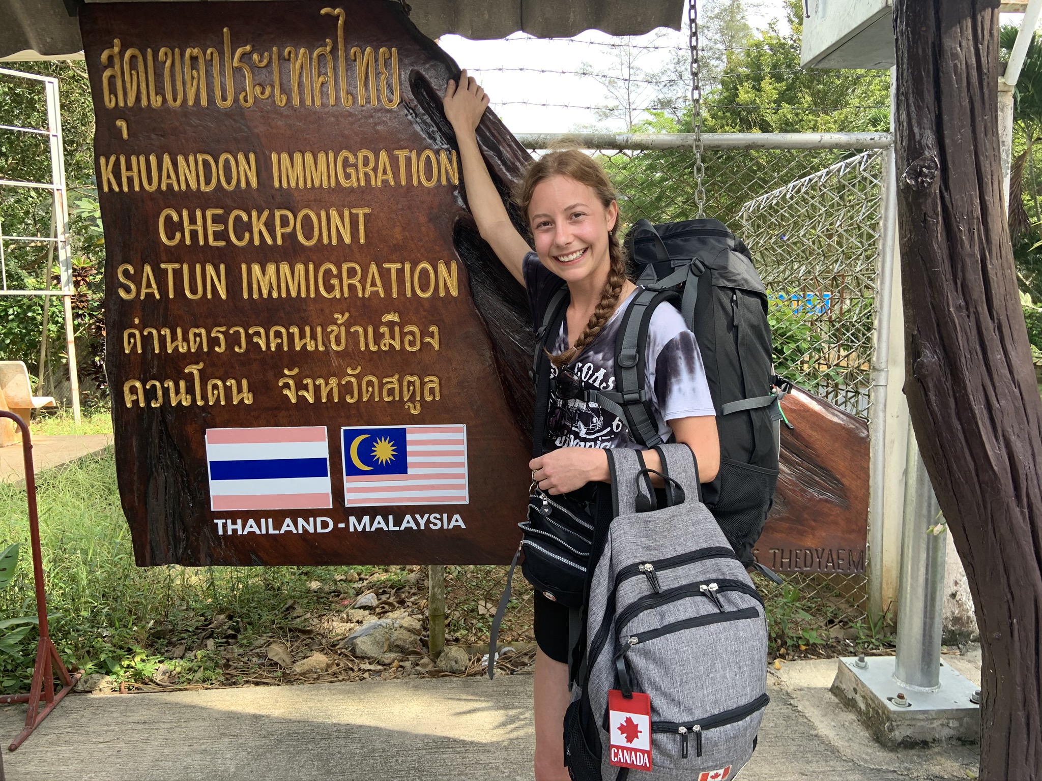 A smiling woman with a long braid of brown hair in a t-shirt and shorts, holding a grey backpack and wearing a large black backpack, next to a sign indicating the immigration checkpoint between Thailand and Malaysia.