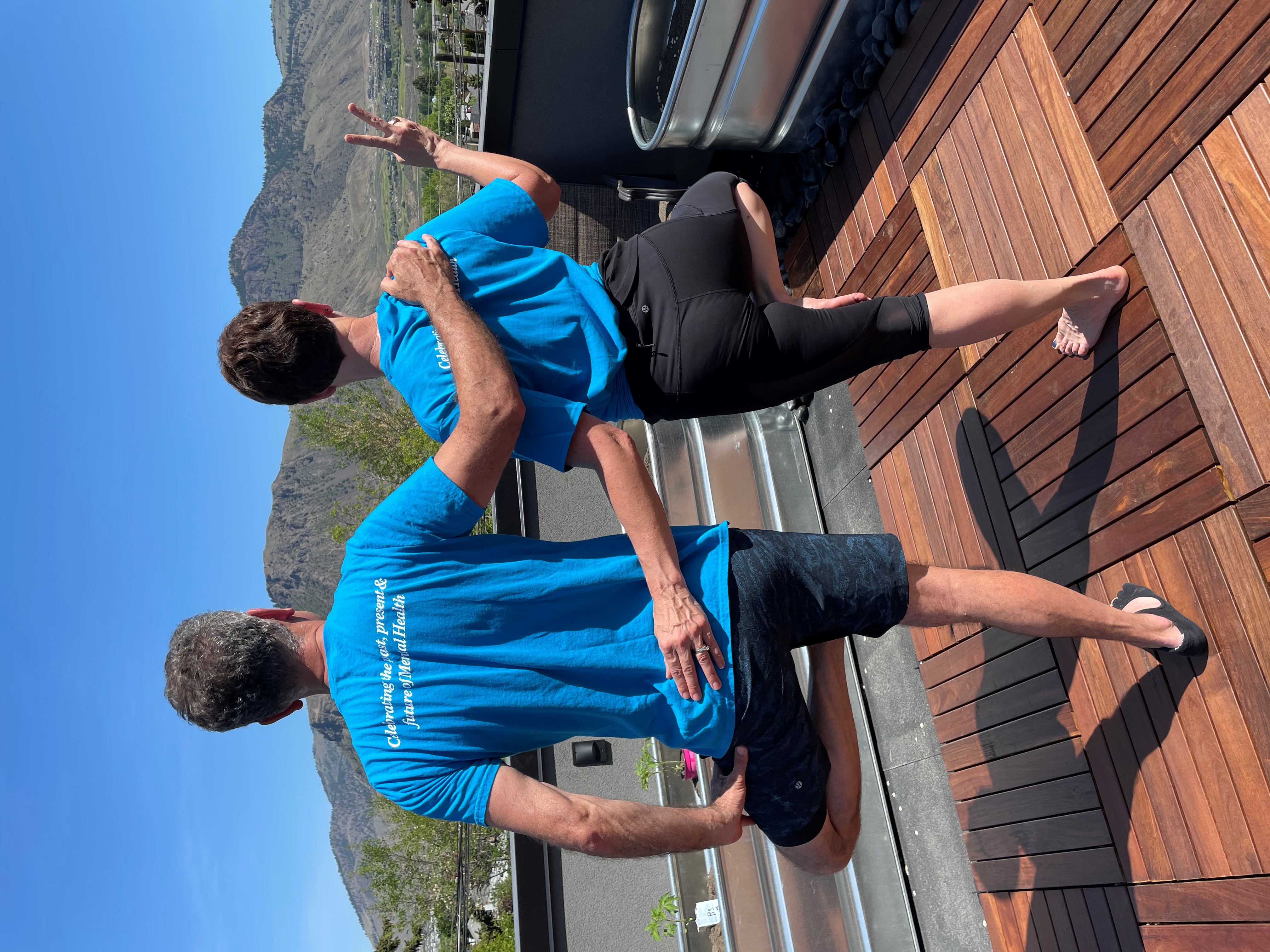 A man (left) and woman in blue shirts and yoga pants standing on one leg with the other leg at a right-angle from the standing leg, with their backs to the camera on a balcony with mountains in the background.
