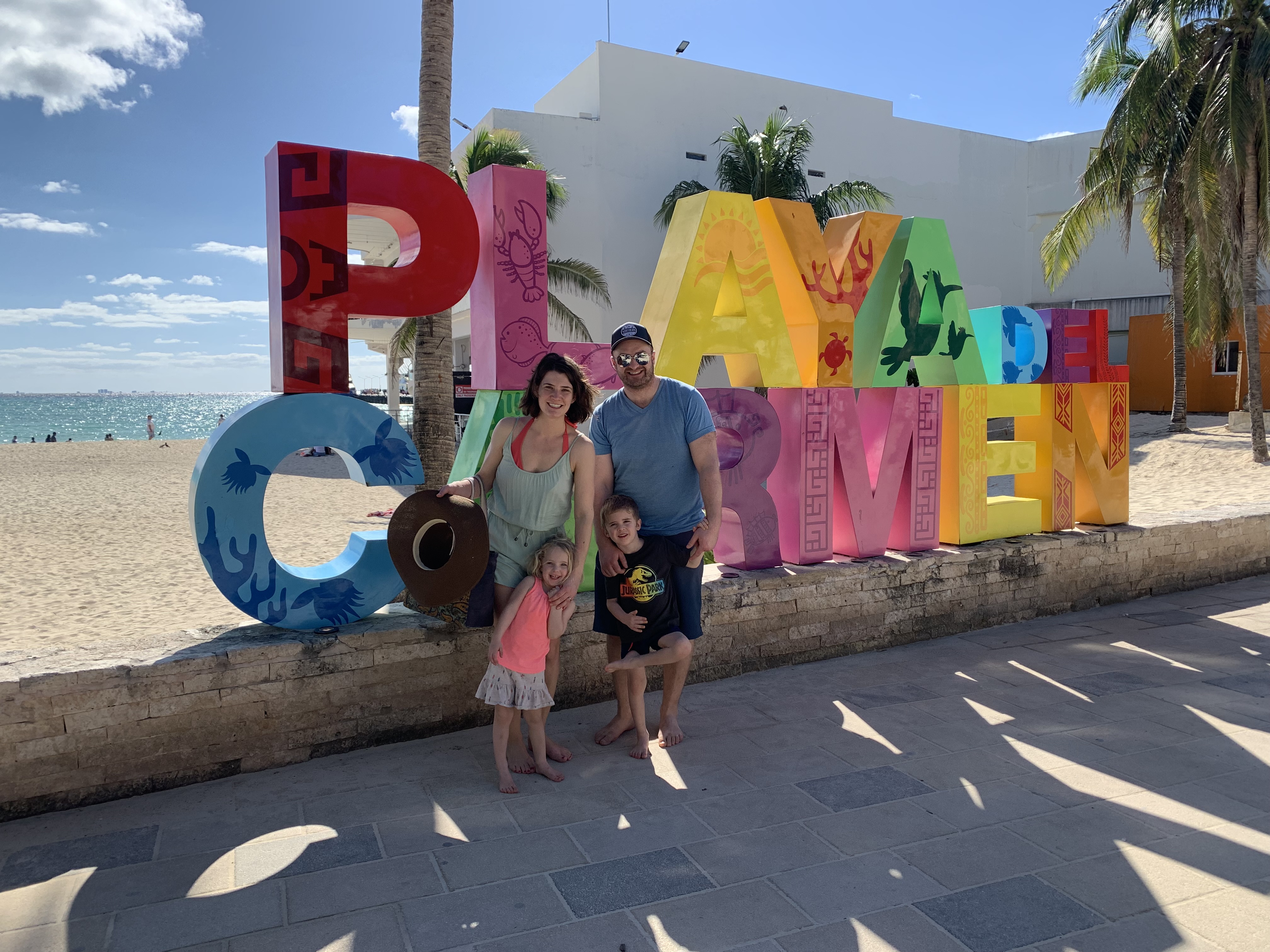 Two adults with two children standing in front of them, all standing in front of a large, colourful sign that reads ‘Playa Del Carmen’ in front of a beach.