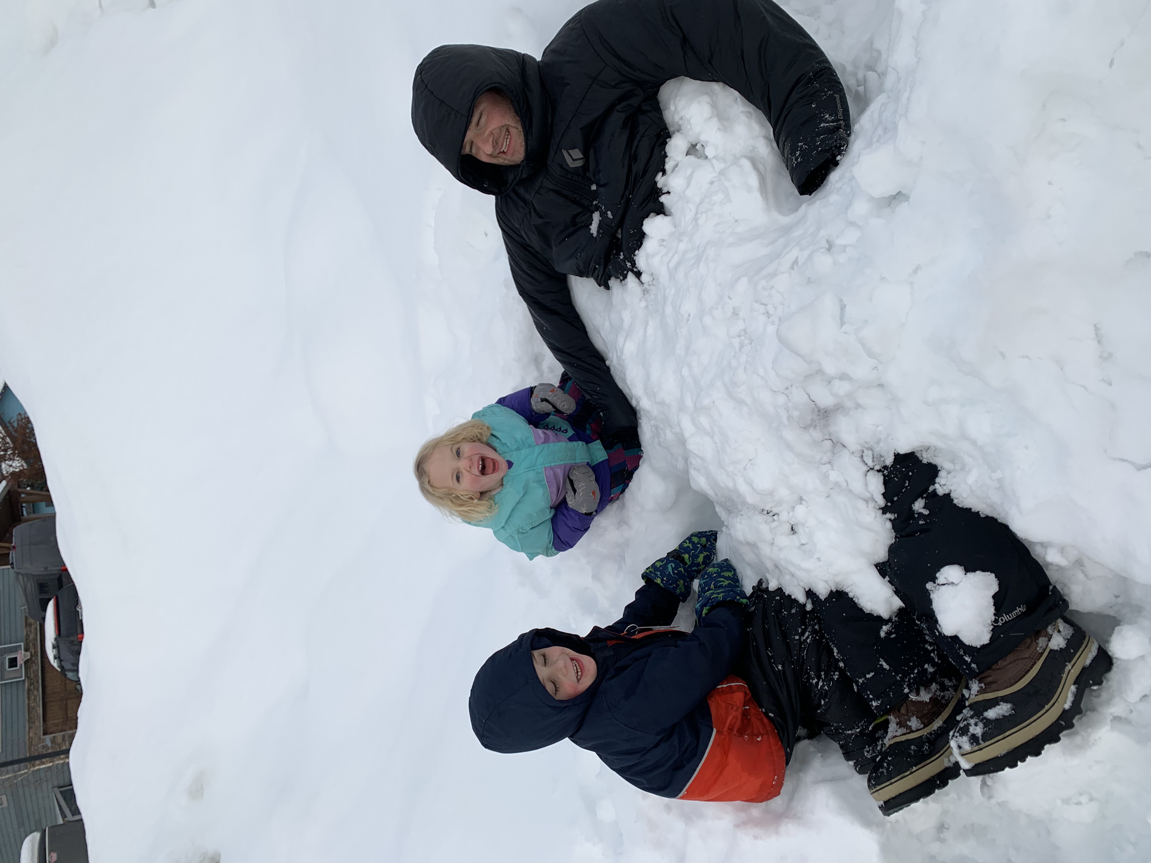 Two children burying an adult in the snow. All are smiling.