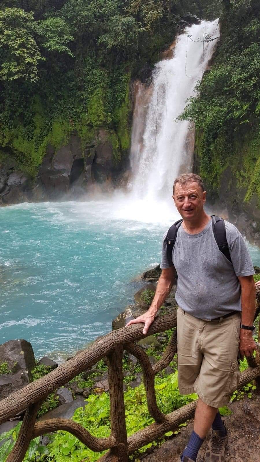 Man wearing a grey t-shirt and backpack holding onto a wooden rail in front of a waterfall that is falling into a light blue body of water.