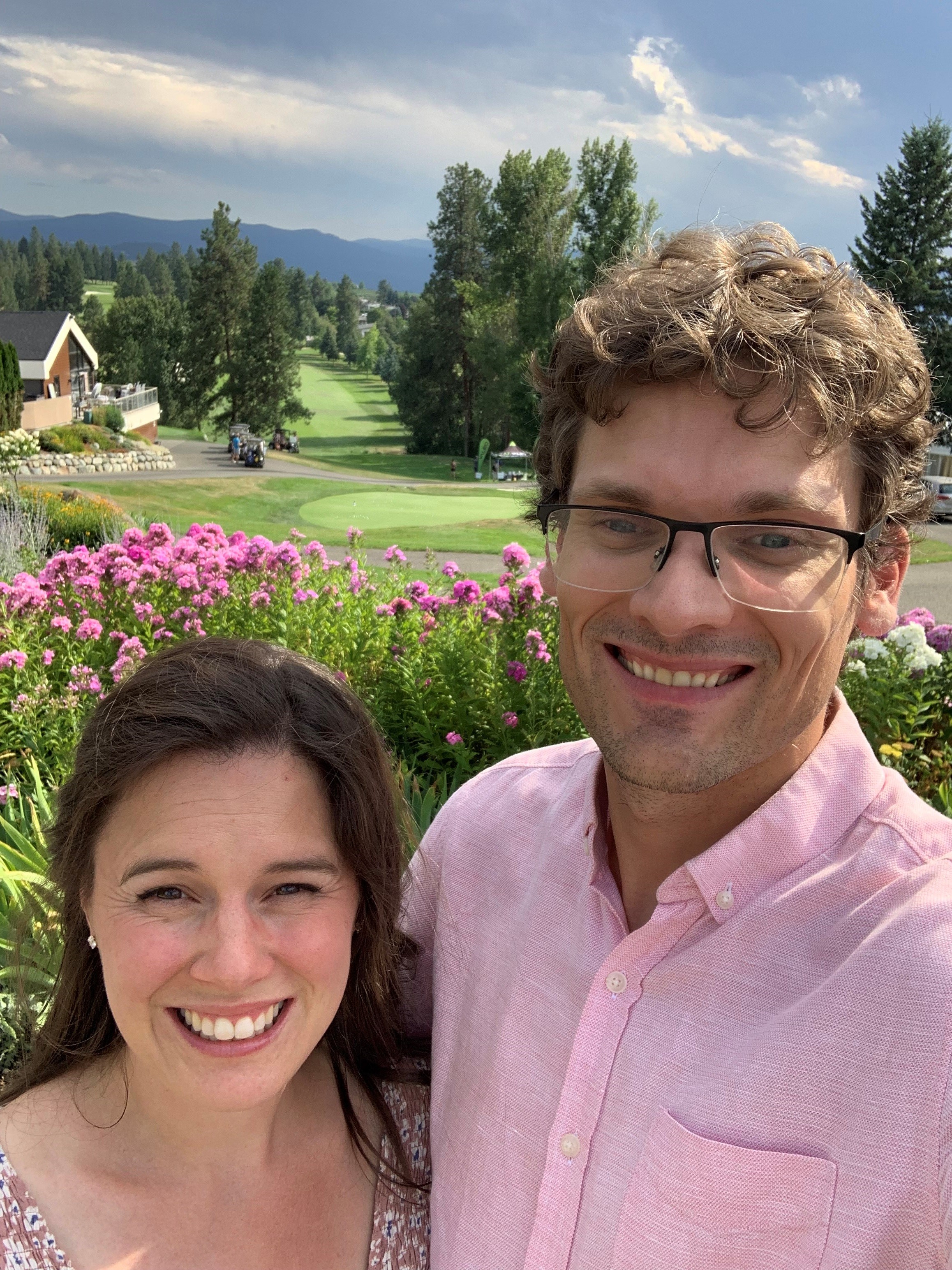 A smiling man and woman with an arm around each other pose for a picture outside with flowers and a golf course in the background.