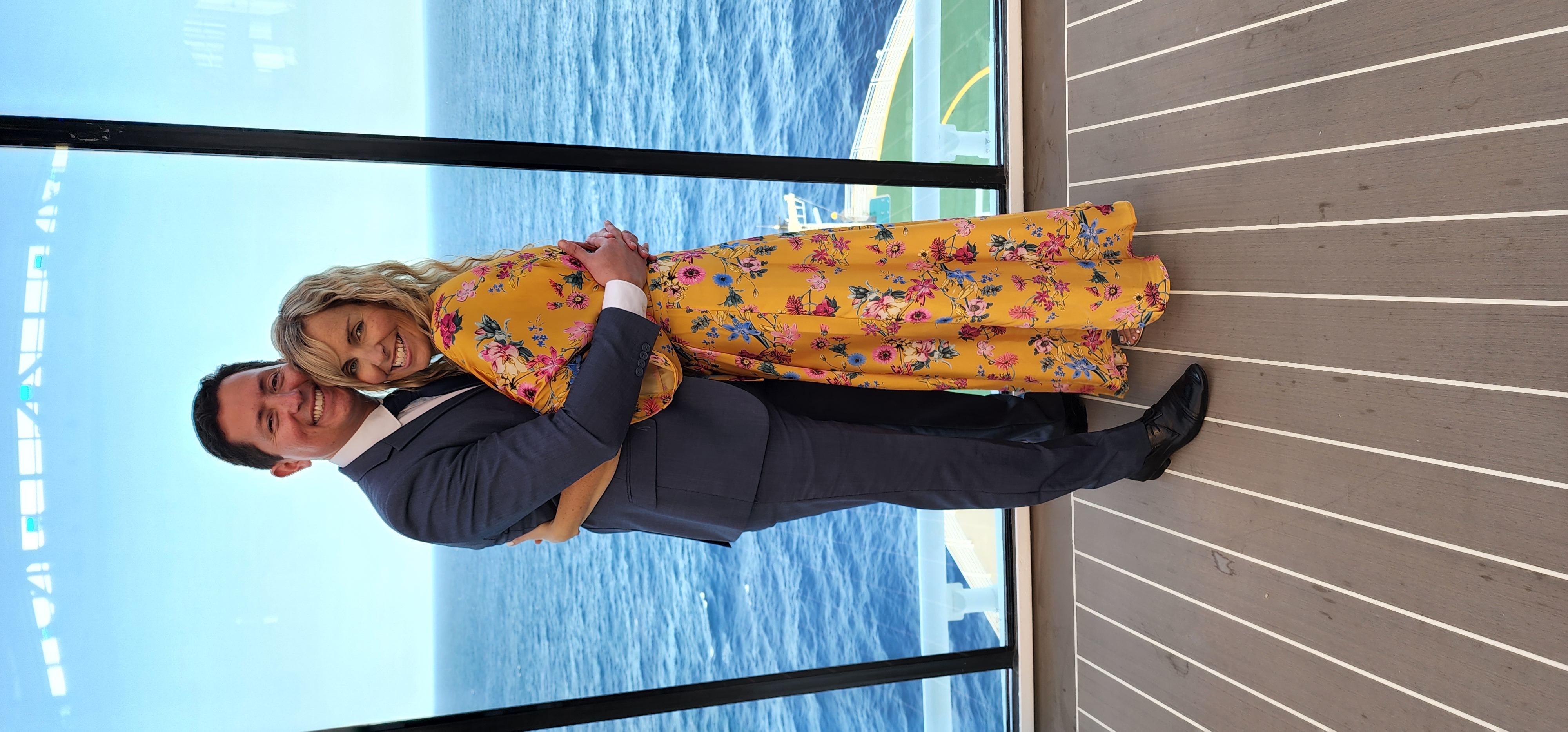 A man and woman in formal evening wear pose for a picture on a cruise ship by a window with the ocean in the background.