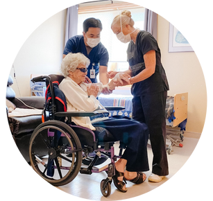 An elderly patient, sitting in a wheelchair in a medical room, has their arm held up by one nurse as another nurse gives them an injection.