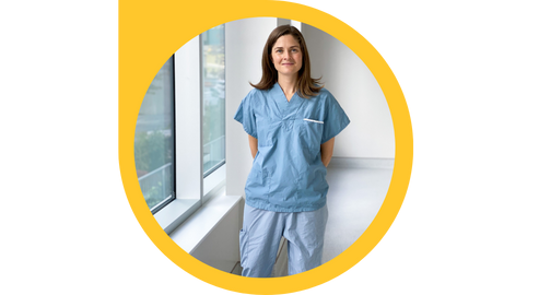 A person wearing blue medical scrubs stands in a hallway next to windows.
