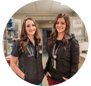 Two people wearing vests and medical gear pose for a picture in a health-care setting.