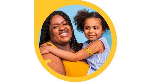 A cheerful adult holds a smiling child who has their arms wrapped around the adult. Both of them have yellow band aids on their arms with various illustrations. 