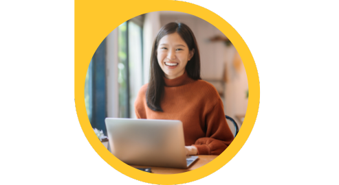 A smiling person with long brown hair wearing an orange turtleneck looks up from a laptop they're typing on while sitting at a desk.