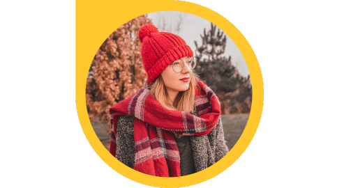 A person wearing glasses and dressed in a jacket, red patterned scarf and red toque stands outside with trees in the background.