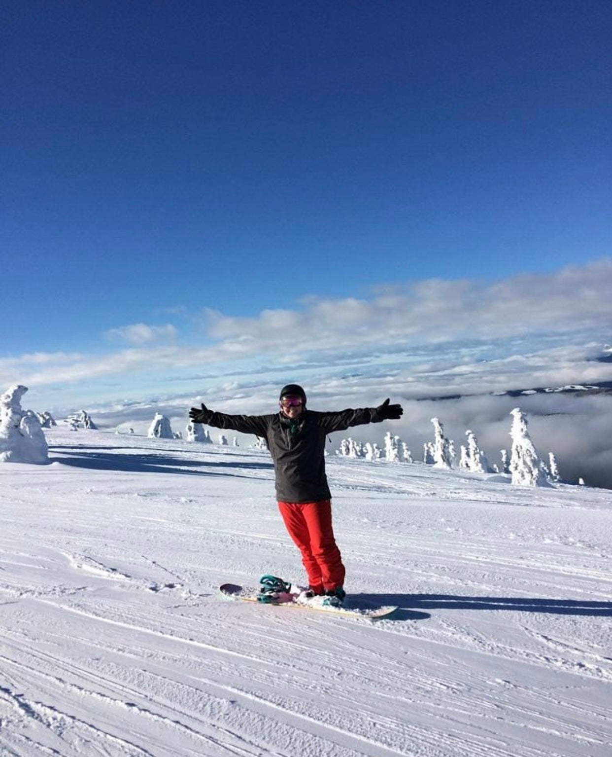 A woman wearing a helmet and bright red snow pants holds out her arms while snowboarding on the top of the mountain while clouds are surrounding the area