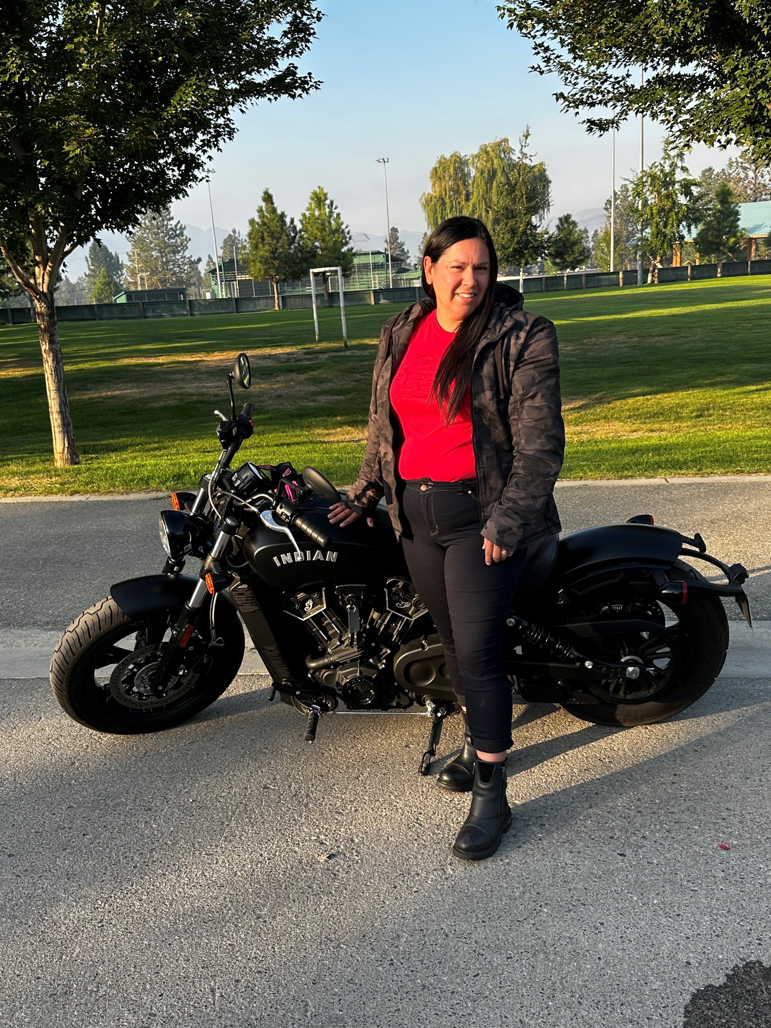 A woman with long brown hair in a bright red shirt leans on an upright motorcycle posed in front of a soccer field