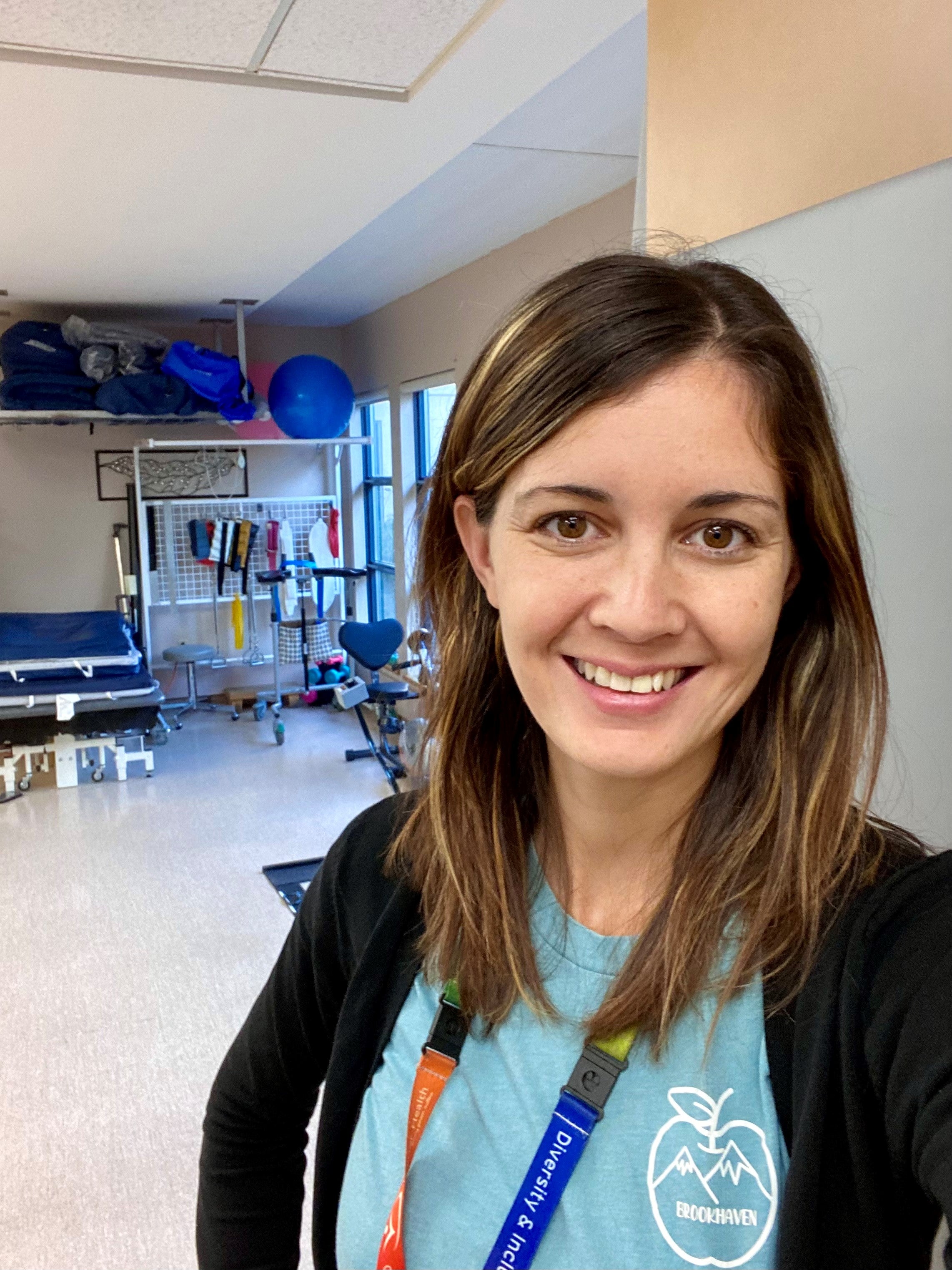 Smiling woman with long brown hair, brown eyes wearing aqua-coloured t-shirt with an apple and mountain logo on it, black sweater and diversity lanyard in a room with exercise equipment