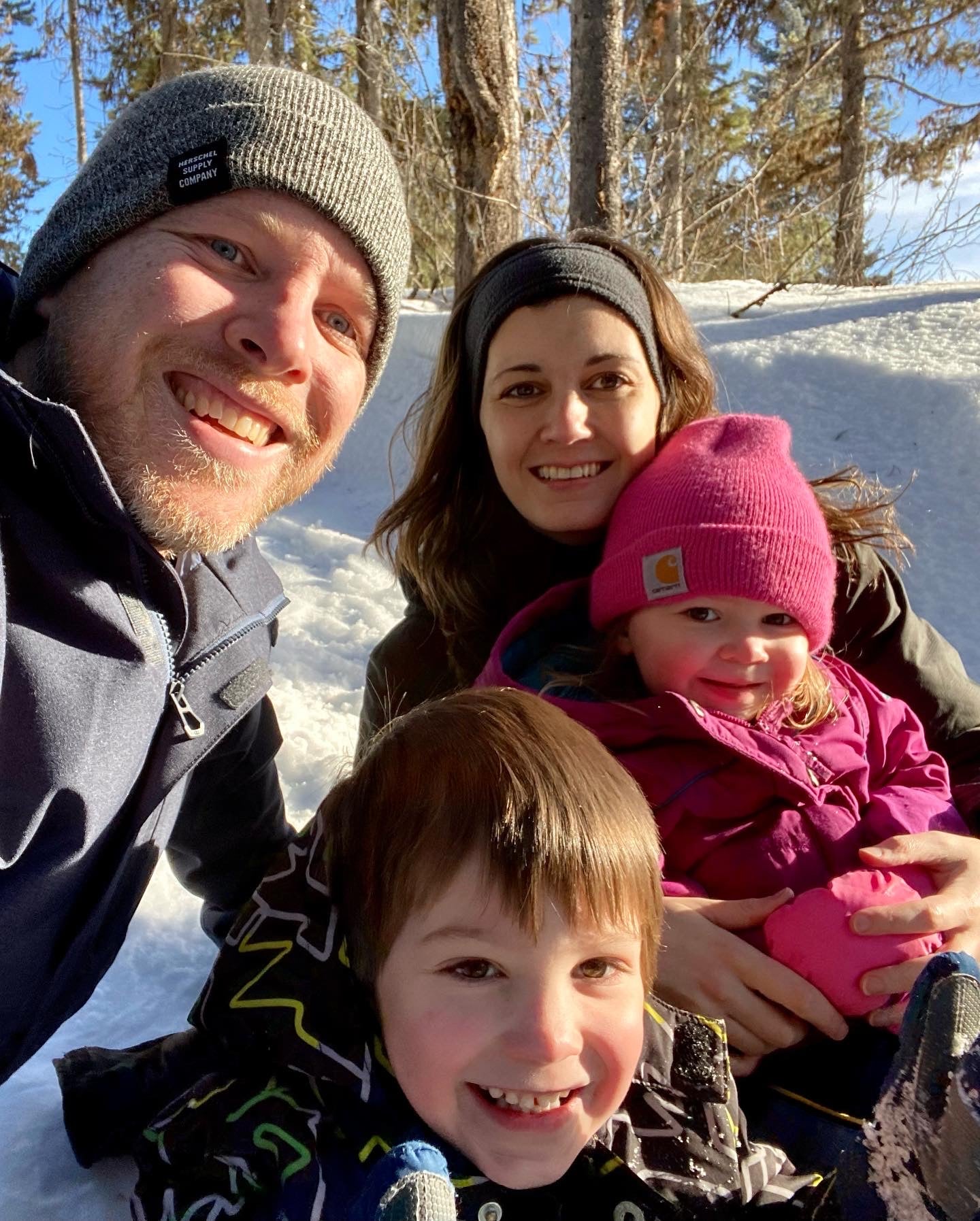 Young smiling family all wearing winter jackets, dad wearing khaki knit hat, mom with long brown hair wearing headband and holding girl daughter with pink toque hat, boy son in front, snow and trees in background