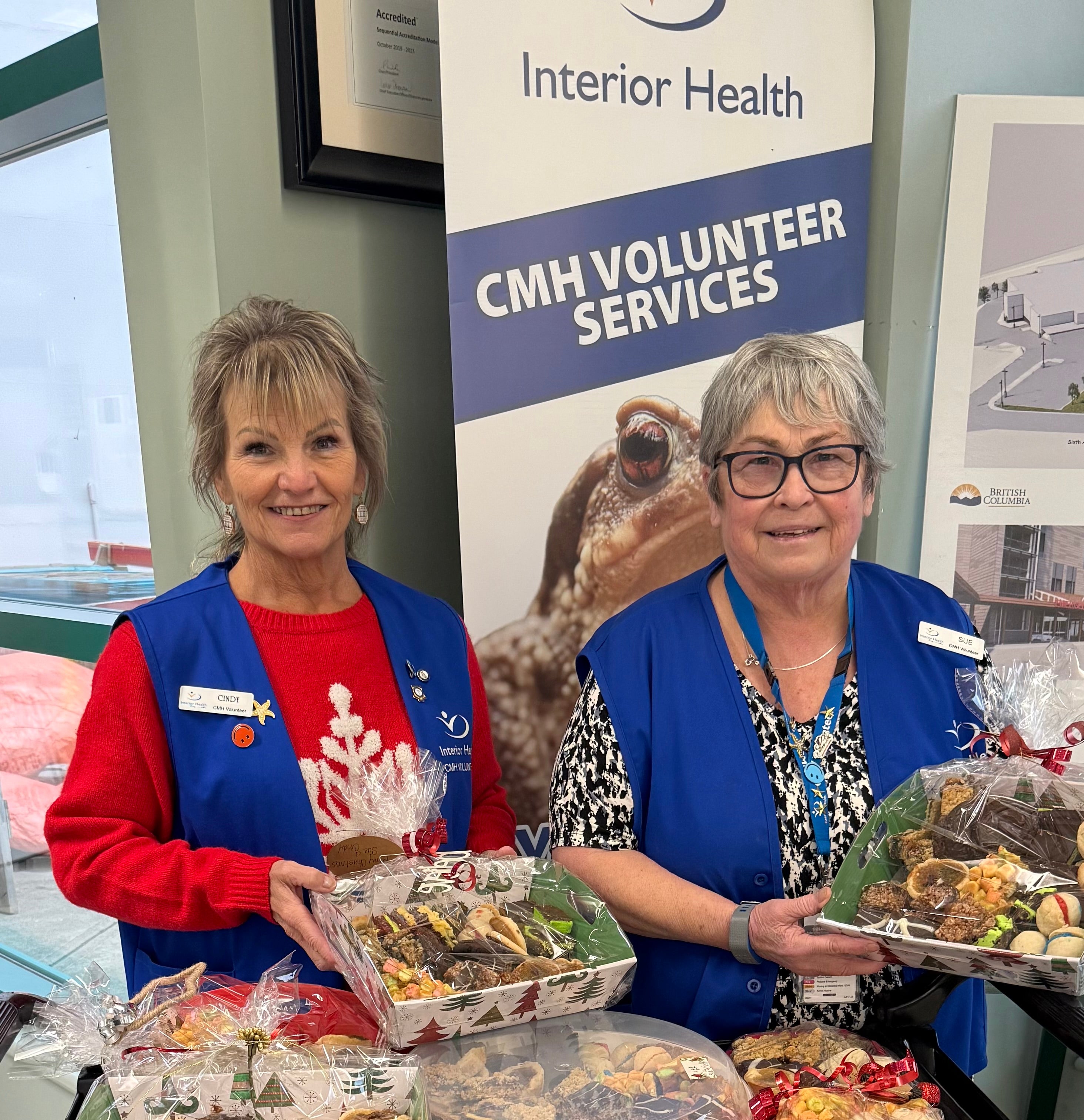 Two people in blue vests both with short grey hair carry plates of food in front of a volunteer services banner.