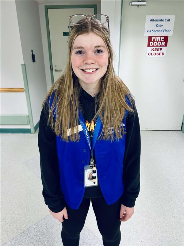 A young person with long blond hair wearing blue vest poses in a hospital hallway.