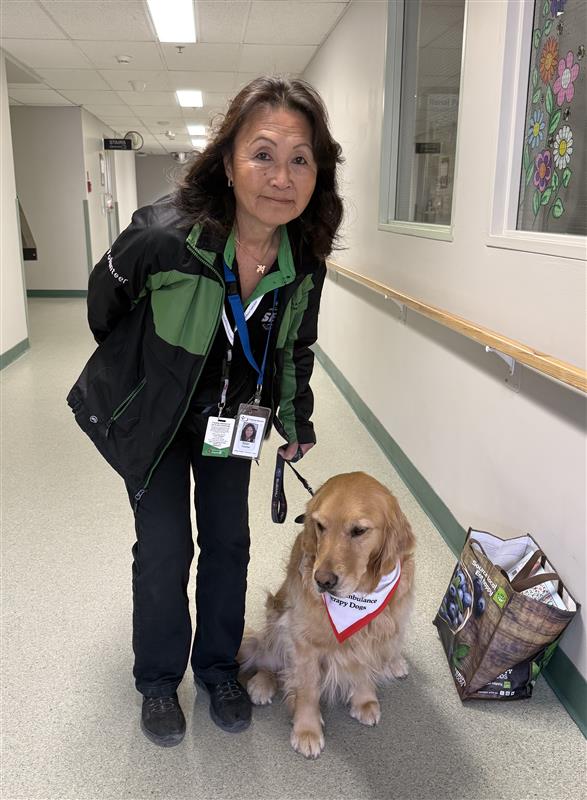 A person wearing a green and black jacket and lanyards holds the leash of a golden retriever in a hospital hallway.