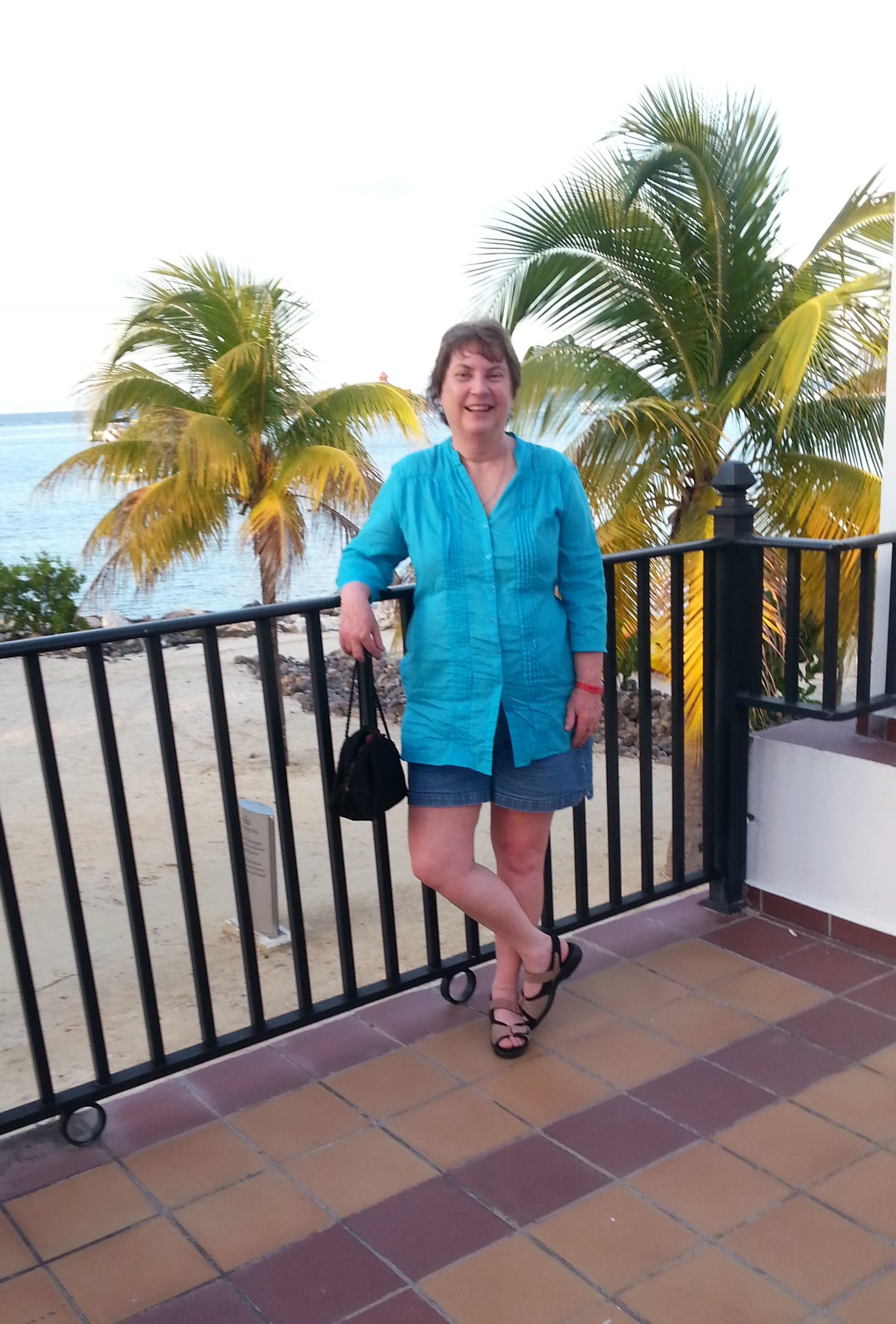 A person with short brown hair in a teal blouse and shorts and sandals stands in front of a black railing at a beach with palm trees