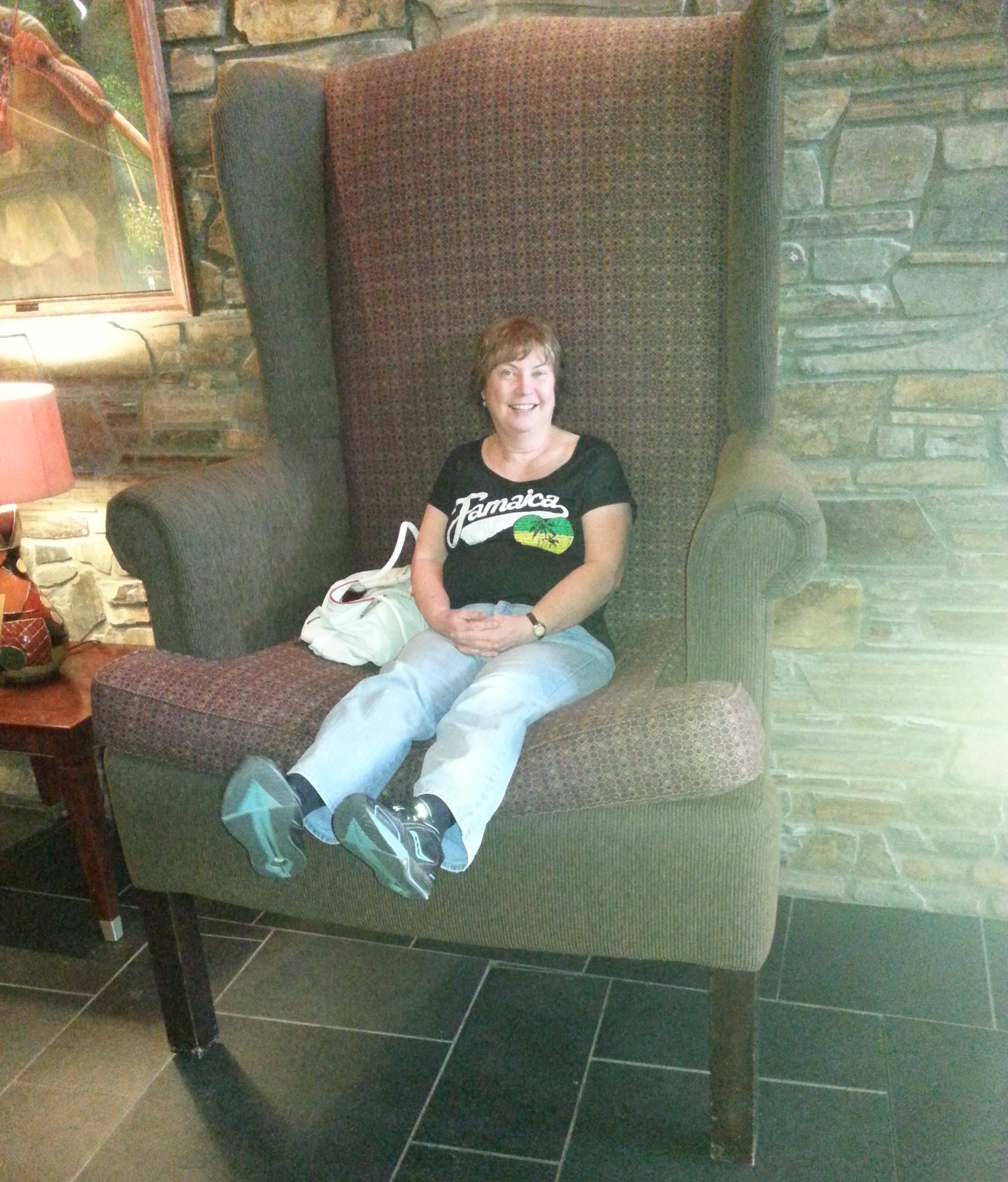 A person in a black t-shirt and blue jeans and sneakers with a white purse sits in an over-sized armwing chair set against a stone wall