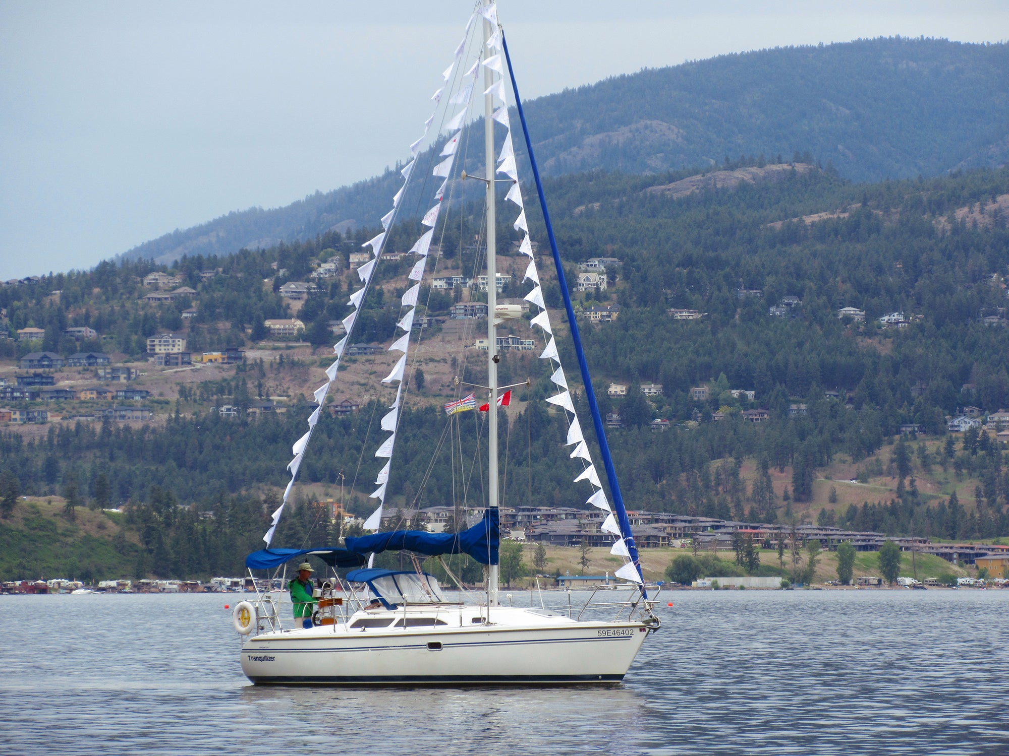 A person wearing a green jacket and Tilley hat pilots a small sailing boat, with forested mountains in the background.
