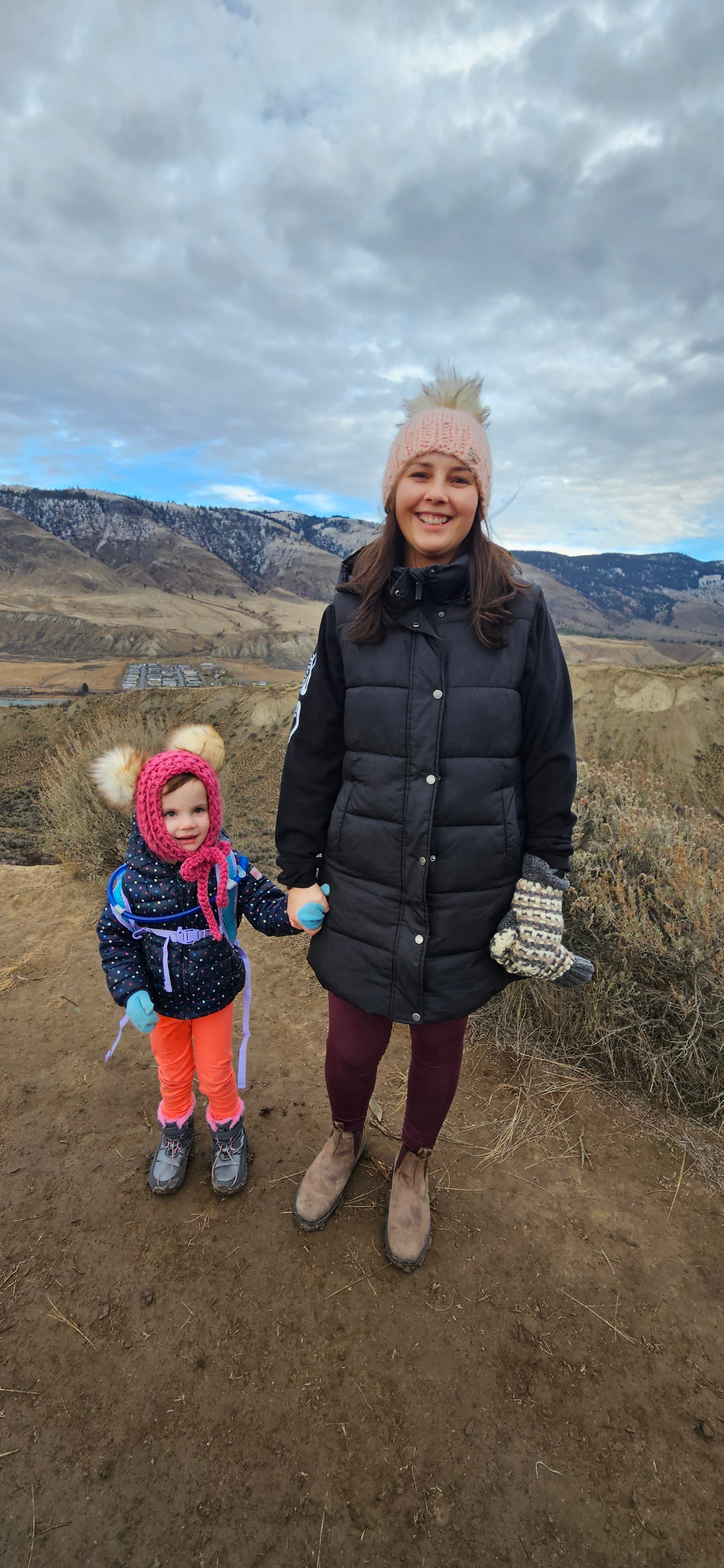 Adult female and small child bundled up in puffy fall jackets wearing crochet toques with fur-style puffs on top and winter gloves standing on hillside with rolling hills, a small community and river in the distance behind them. 