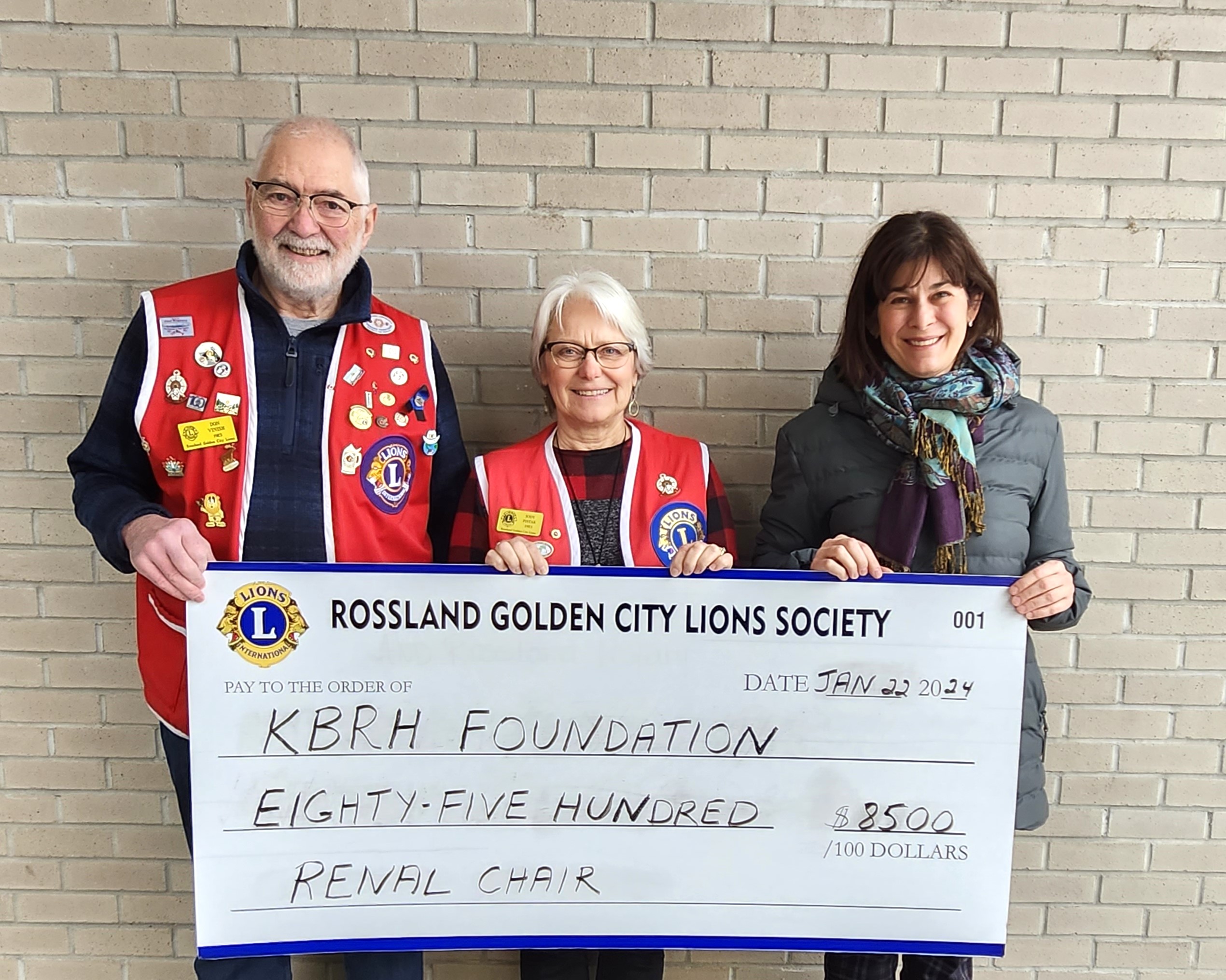 Three people, two of them wearing red vests full of pins, hold a cheque from the Rossland Golden City Lions Society to the KBRH Foundation for $8,500.