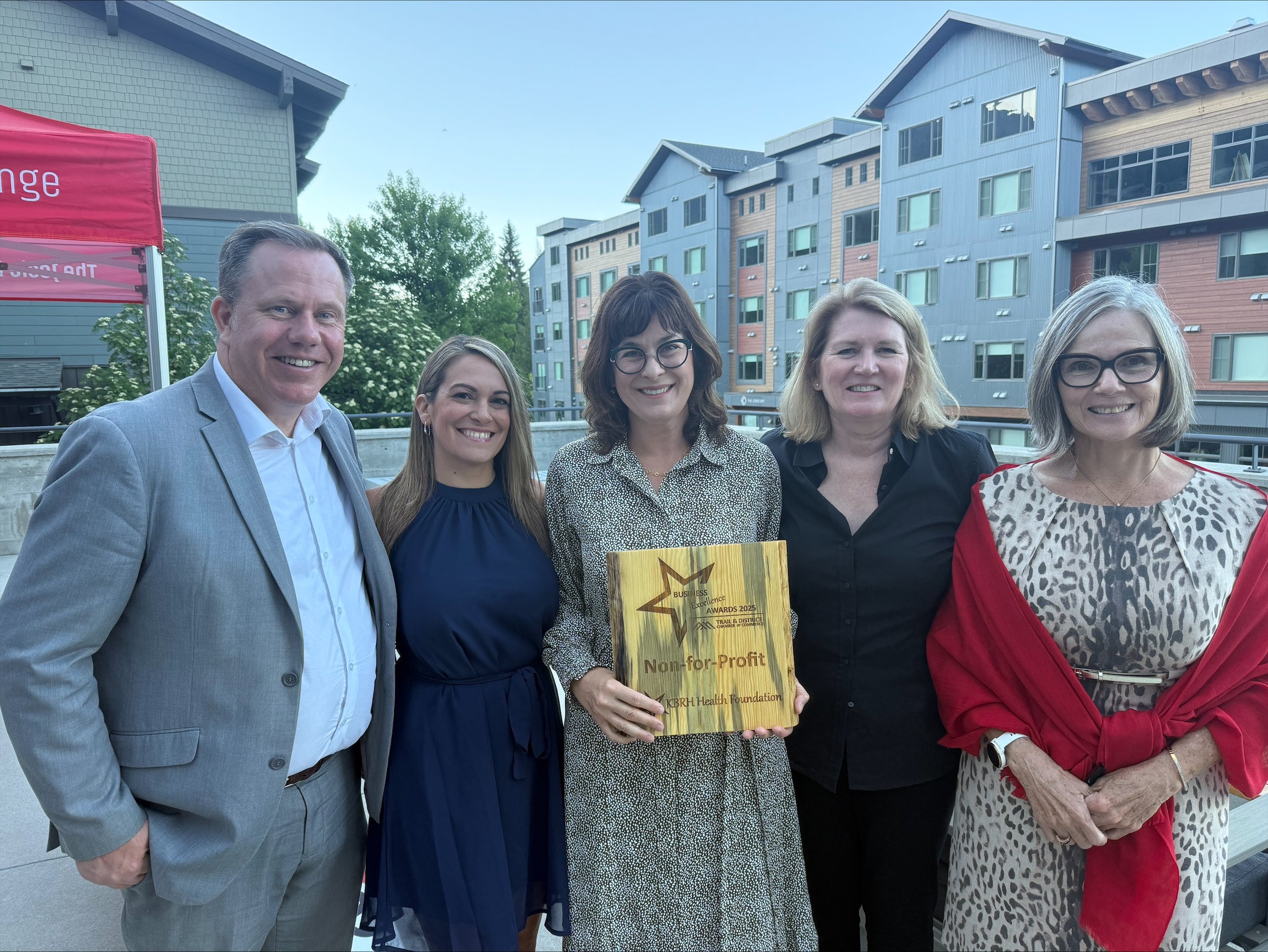 A group of five people in formal business attire pose  on a rooftop patio for a photo with a wooden award plaque that says Non-for-Profit.