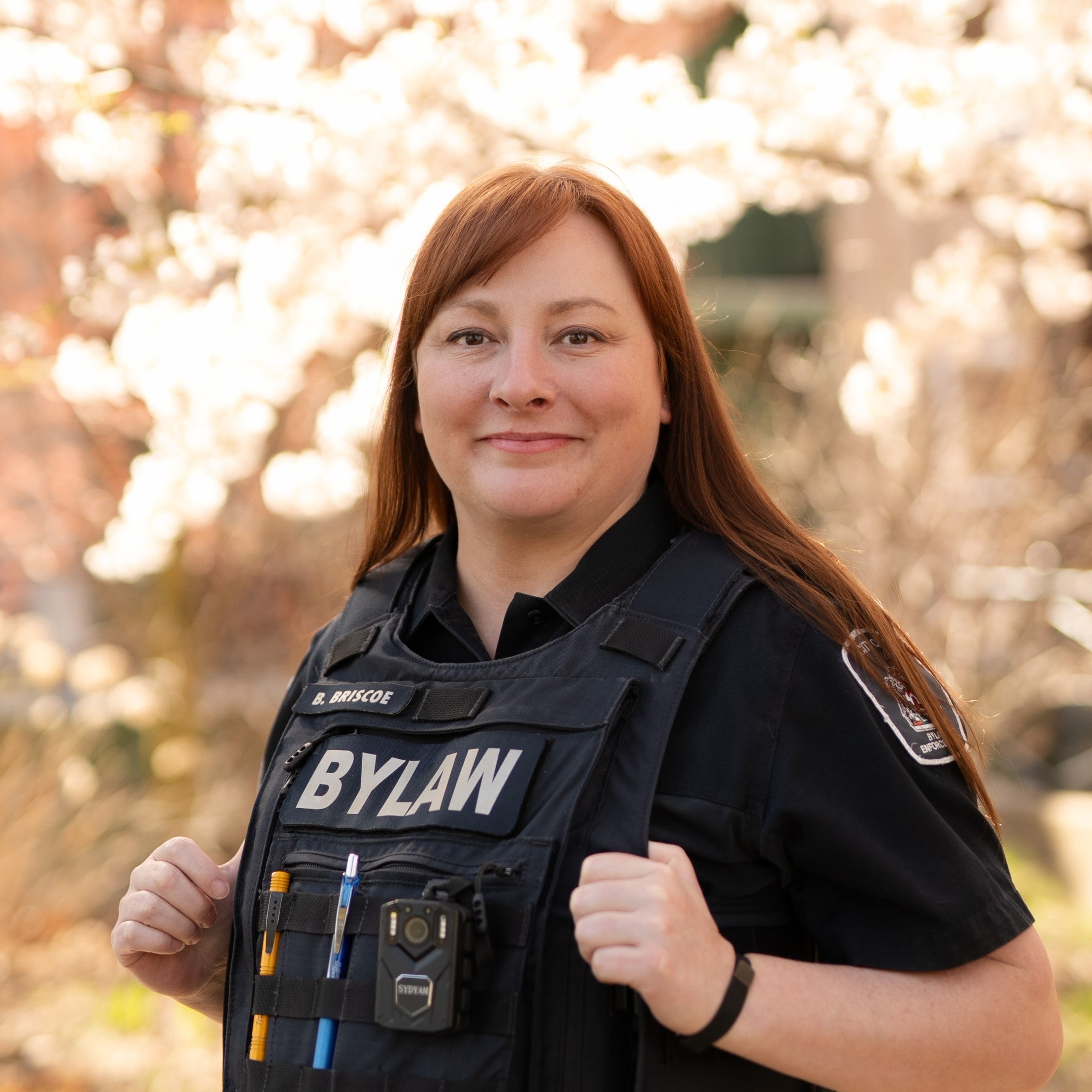A person with long reddish hair standing among cherry blossoms. They have their thumbs hooked into a vest that says Bylaw.