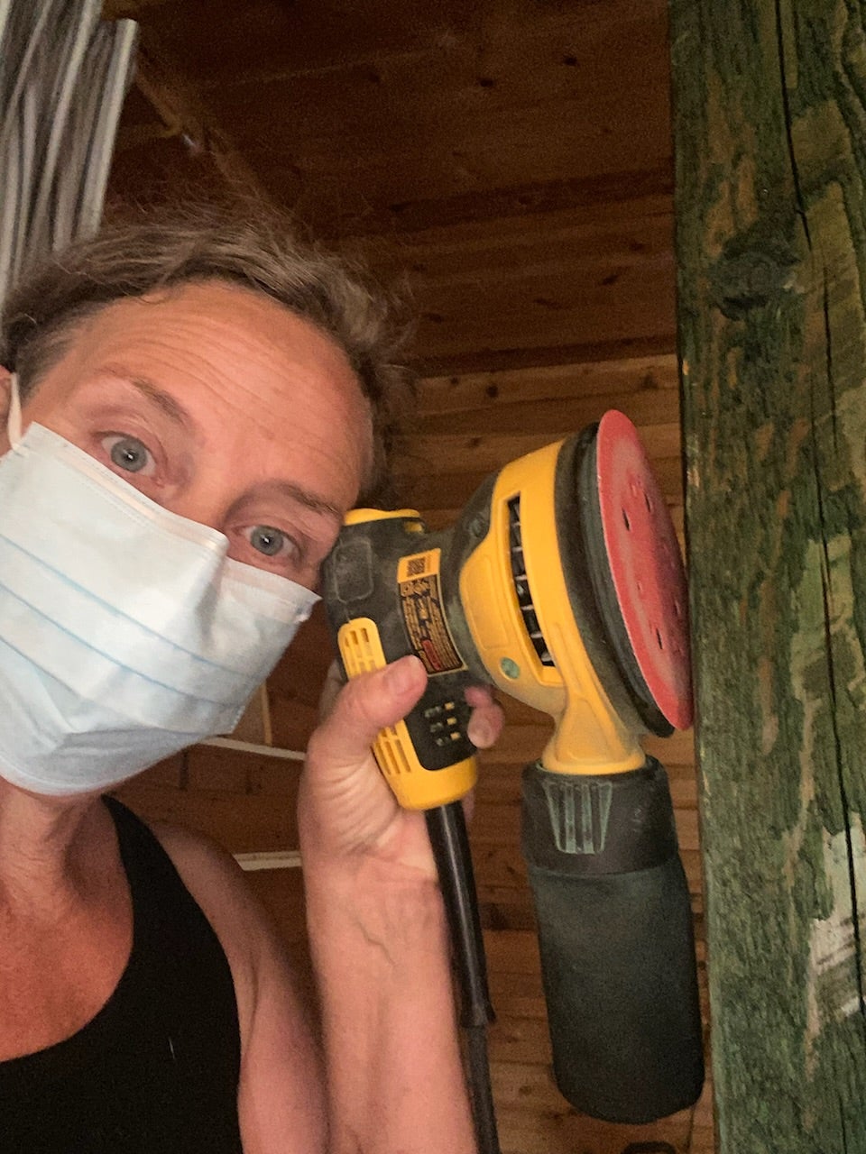 A person wearing a mask and blank tank top holds up a sander against a wood beam.