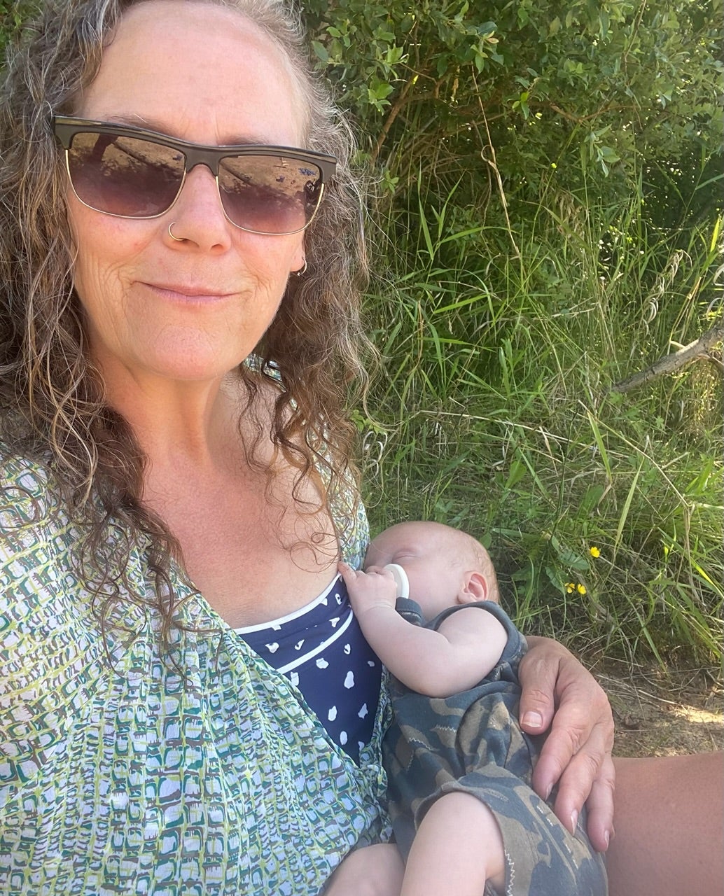 A person with long curly grey hair, sunglasses and green blouse holds a baby in a green camo onesie.