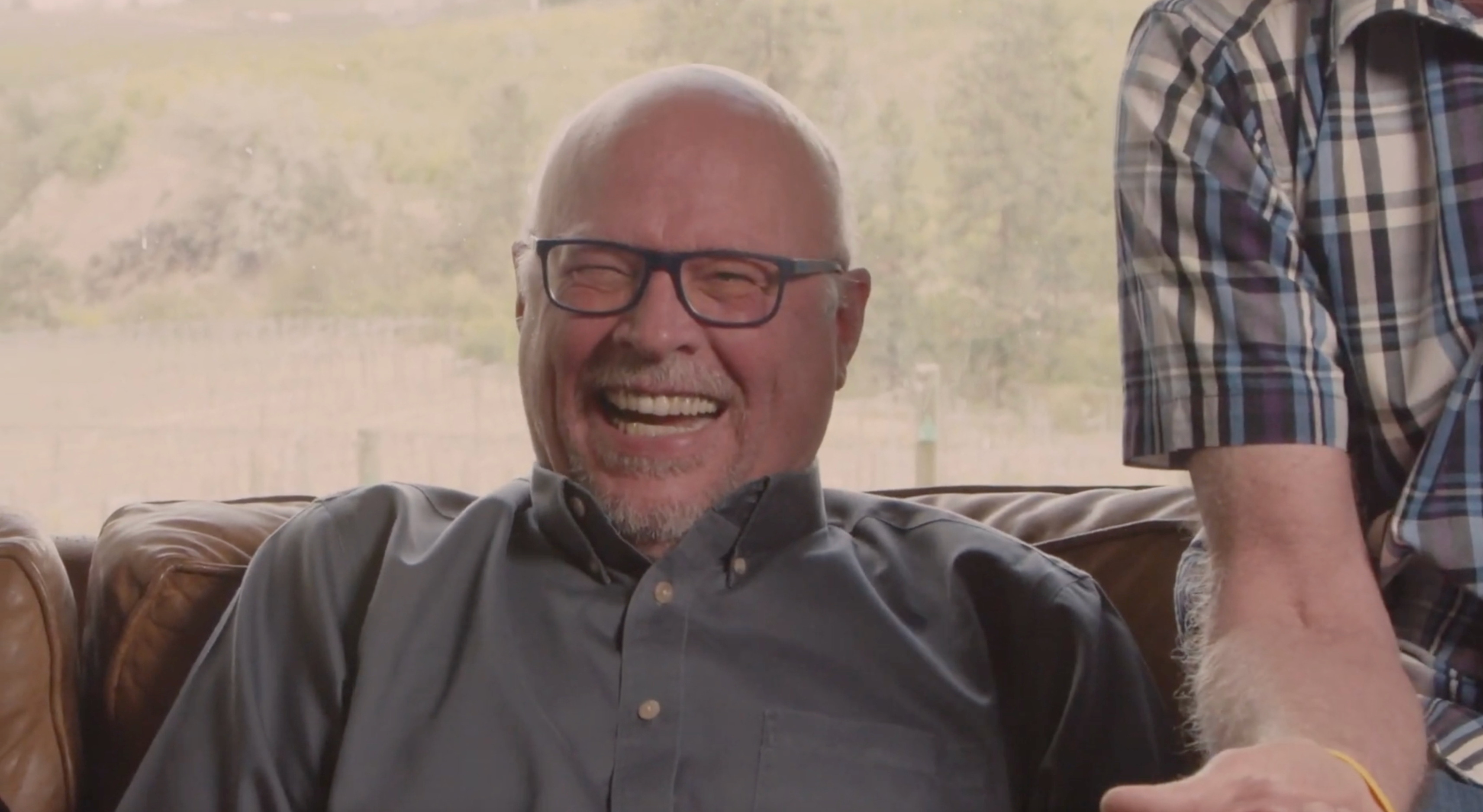 A laughing older man with glasses and thinning hair and wearing a grey button shirt sits on a brown couch.