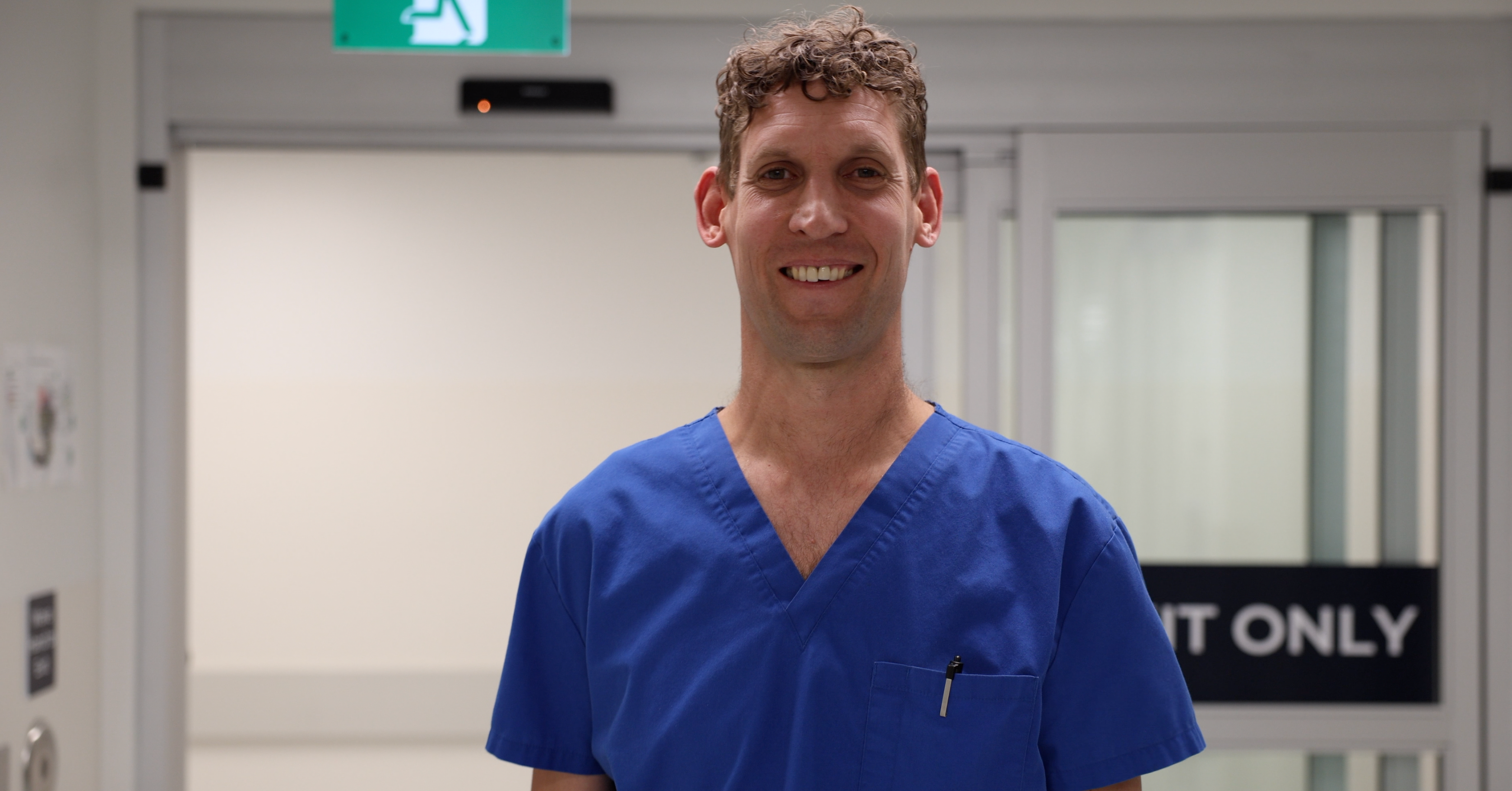 A smiling man with curly brown hair and wearing blue medical clothes stands in front of sliding doors at a hospital.