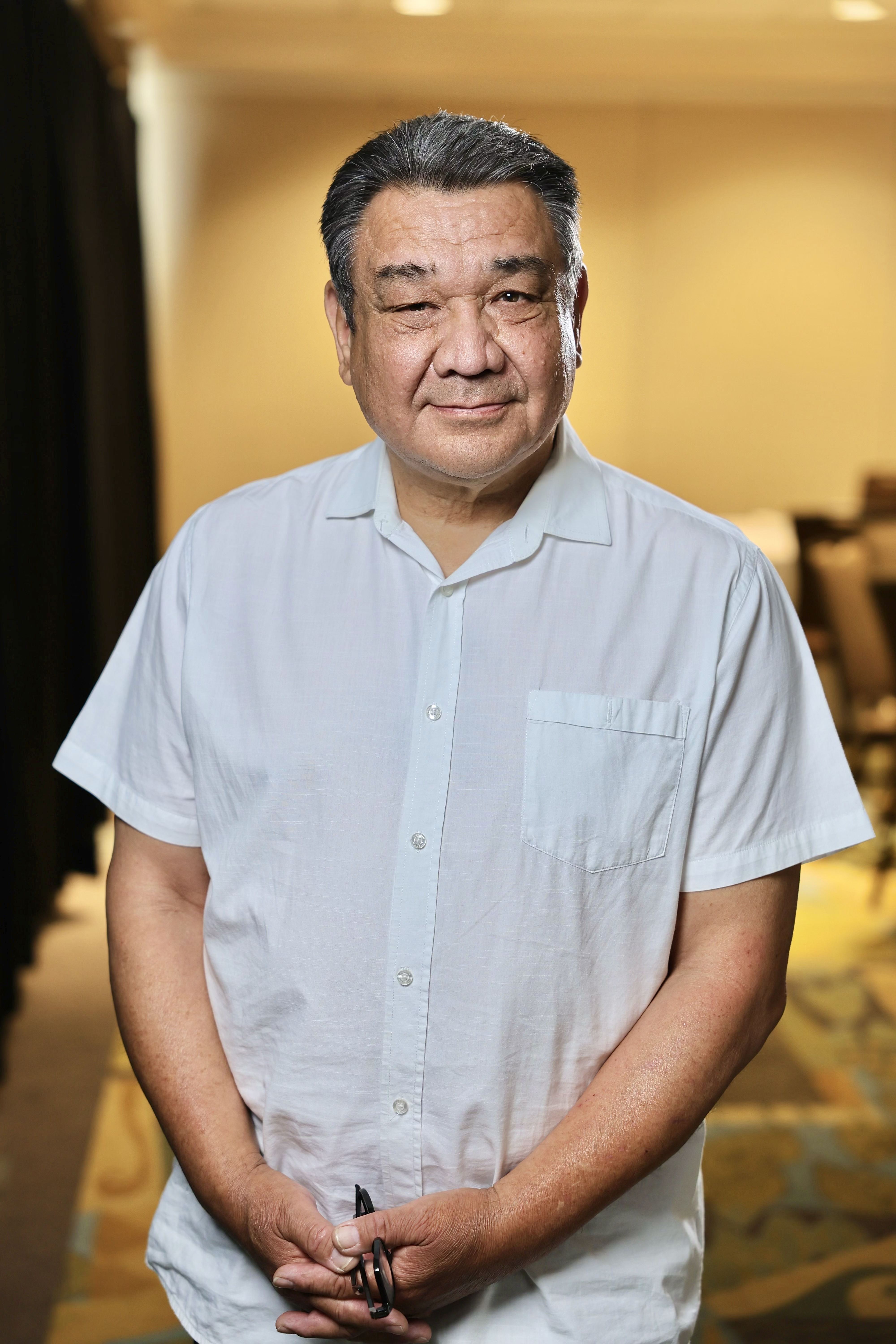 A middle aged man with black and silver hair, who is wearing a light blue button up shirt 