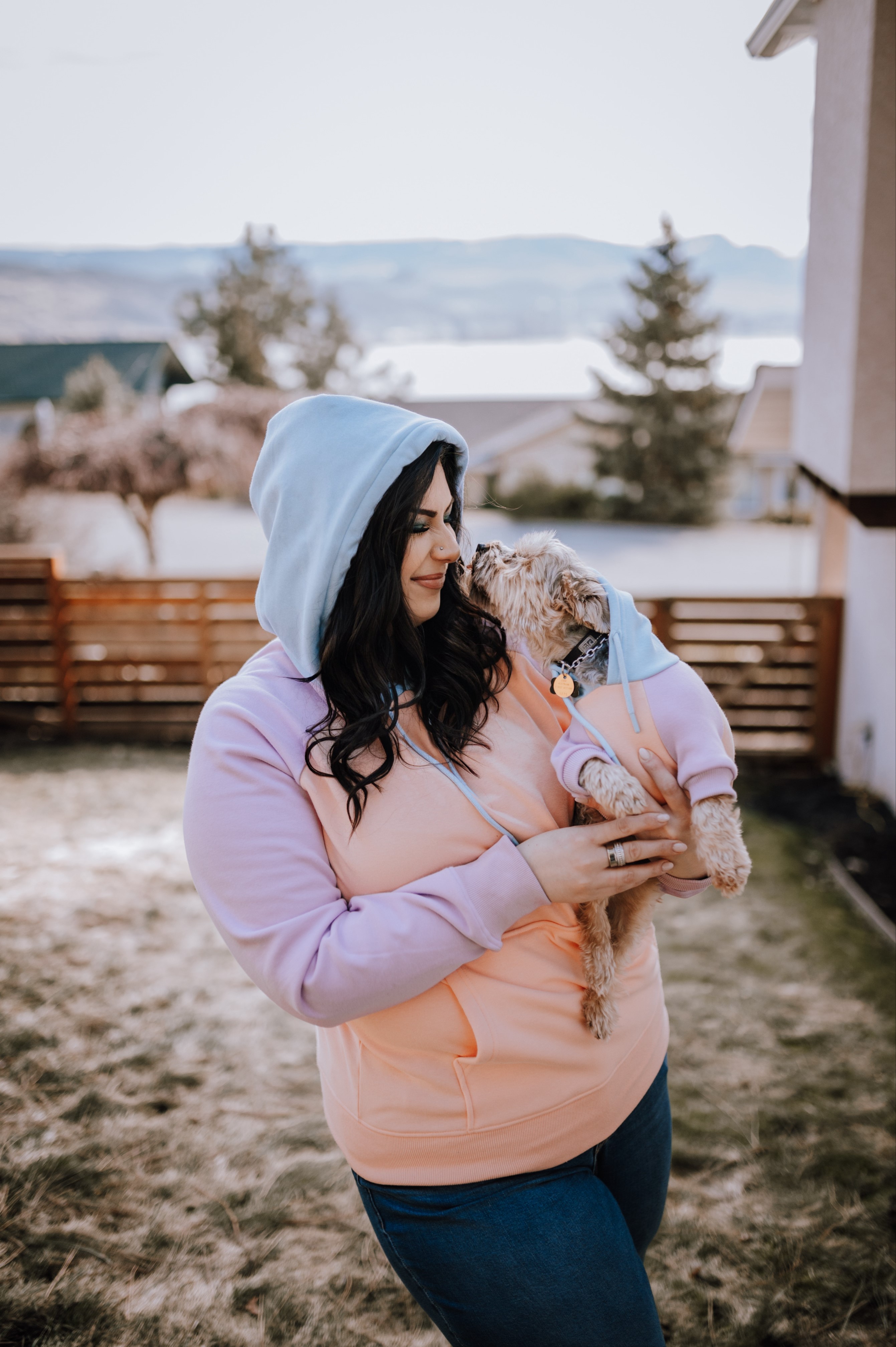 Woman holding a small dog that is kissing her face, both are wearing matching sweatshirts.