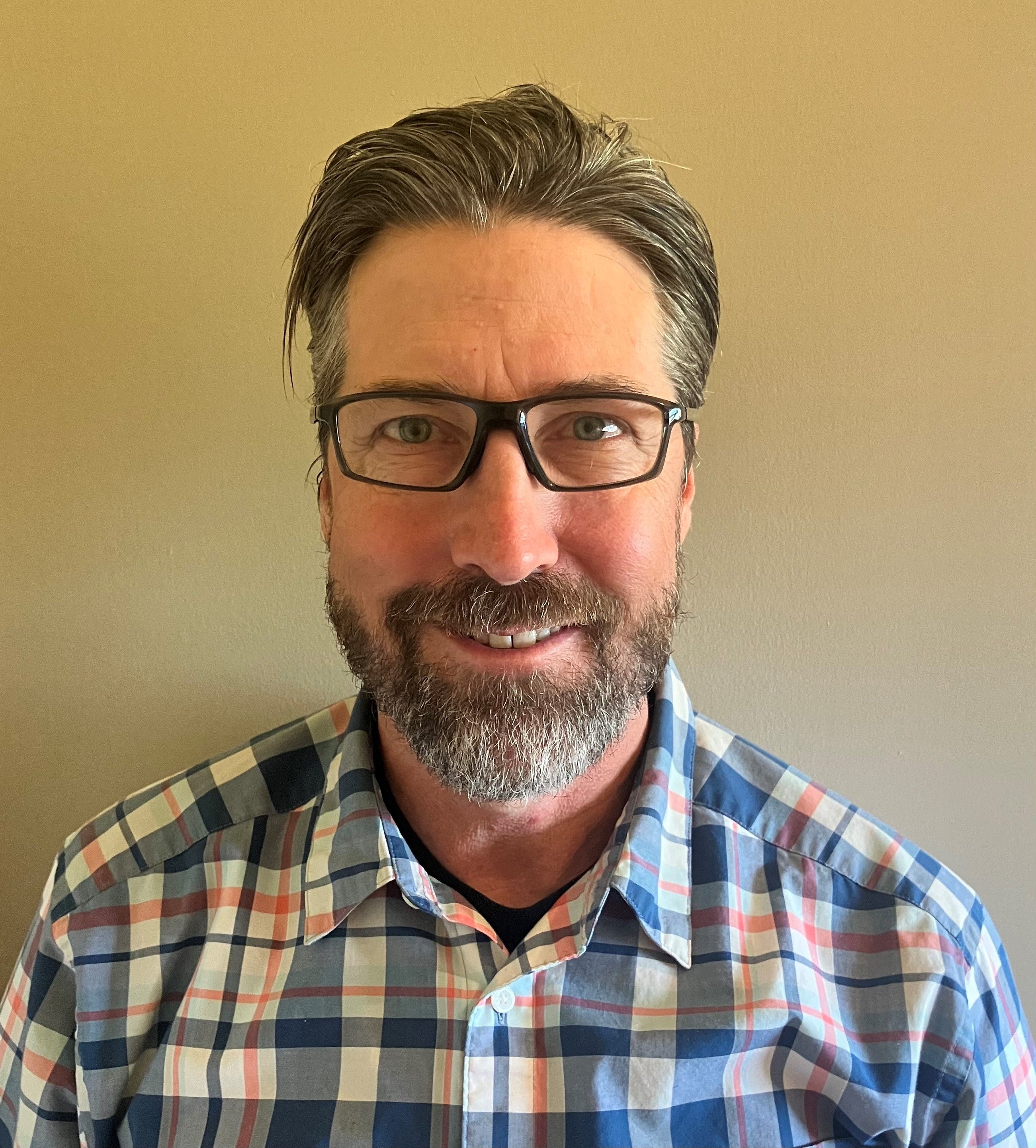A profile shot of a man wearing glasses with wavy dark hair and beard in a multi-coloured checkered shirt.